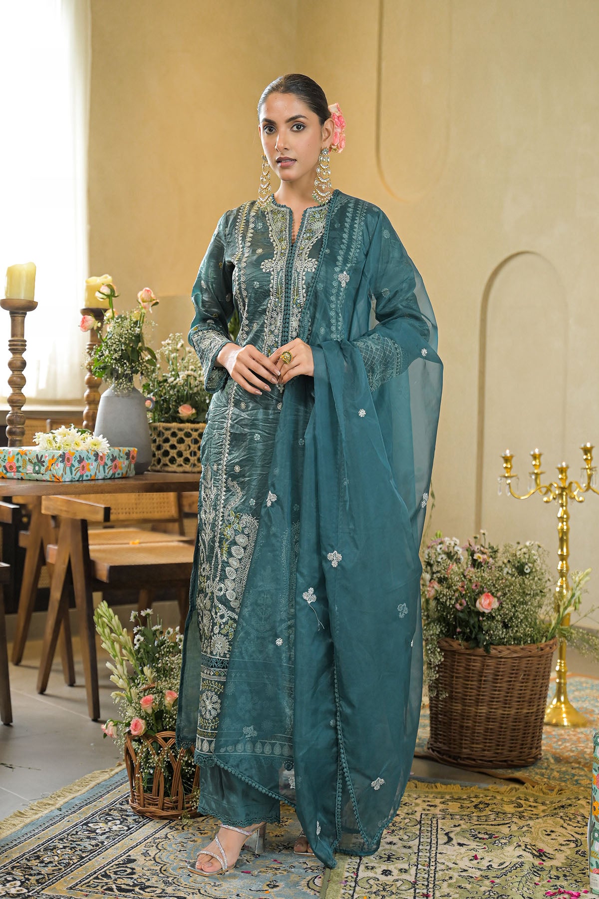 Woman in a teal traditional outfit standing in a decorated room with flowers and candles.