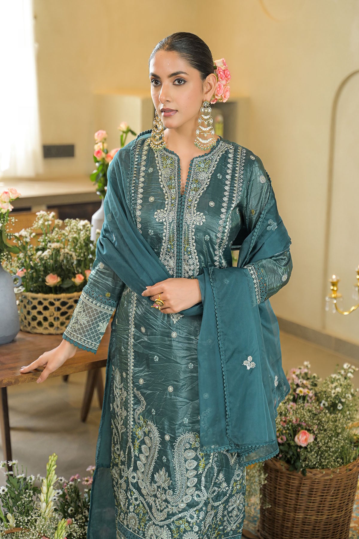 Woman in a teal embroidered outfit standing in a decorated room with flowers.