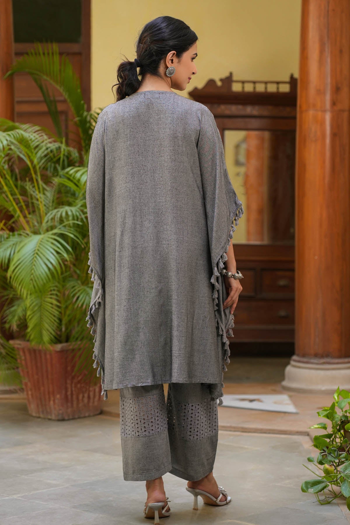 Woman in a gray outfit standing in an indoor setting with plants and wooden columns.