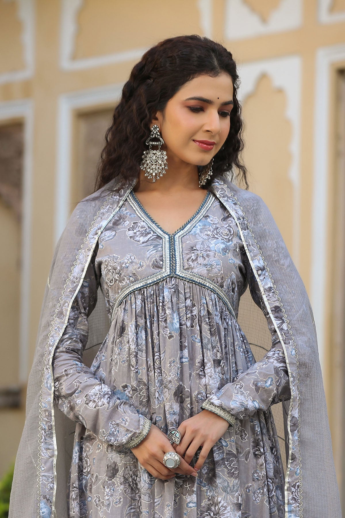 Woman in a silver traditional outfit with intricate designs against a beige wall.