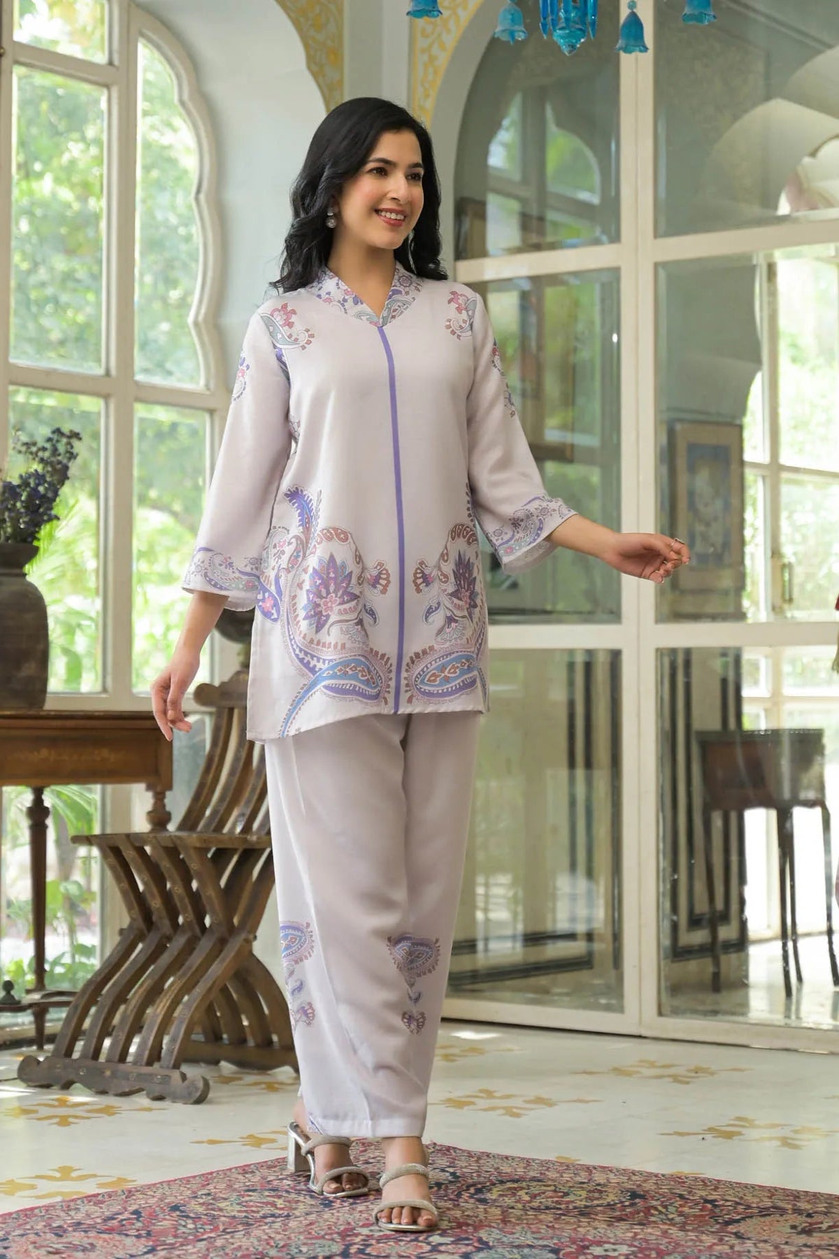 Woman in a light-colored embroidered outfit standing in a room with large windows.