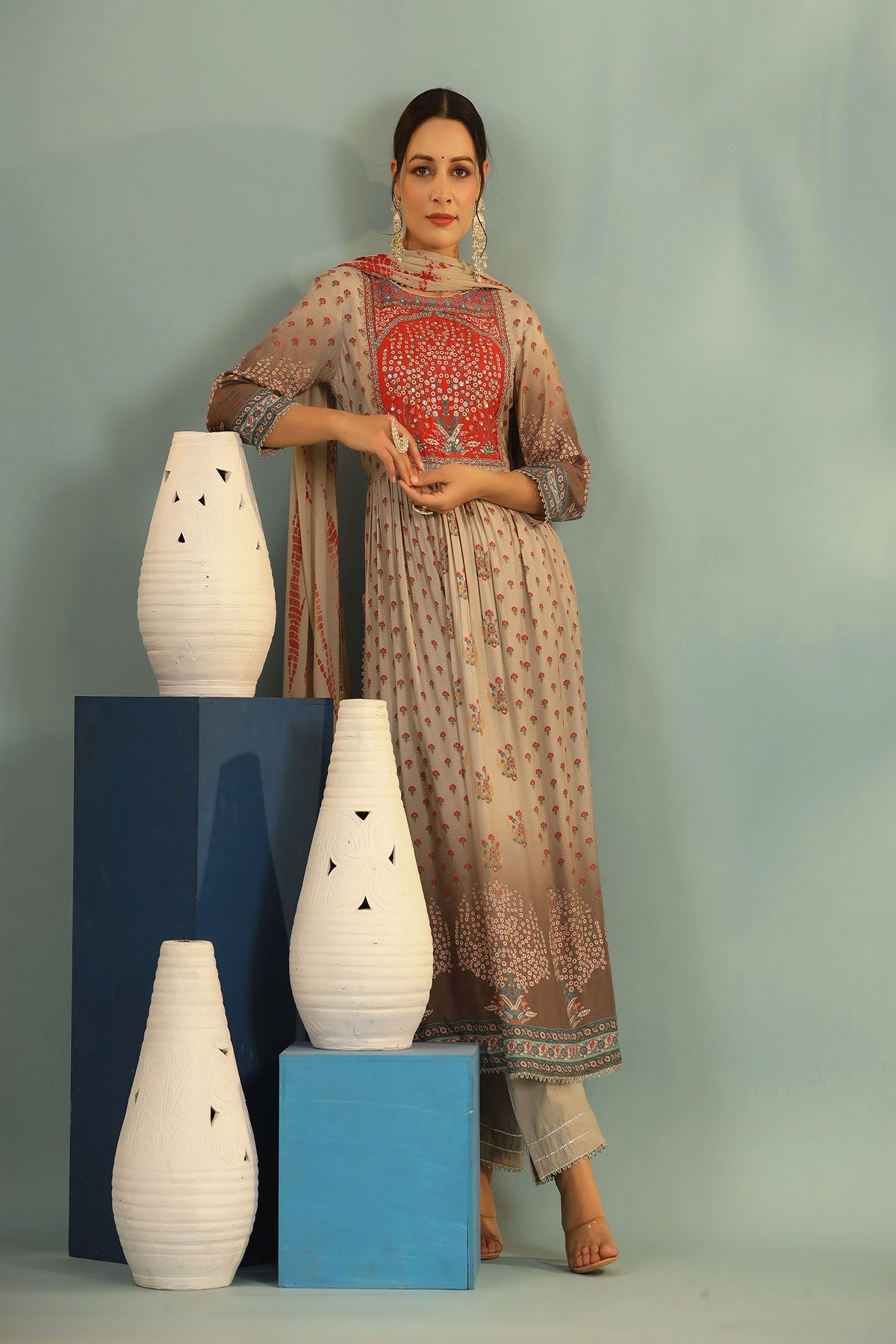 Woman in traditional outfit holding a vase with decorative patterns, standing next to white vases on blue stands against a gray background.
