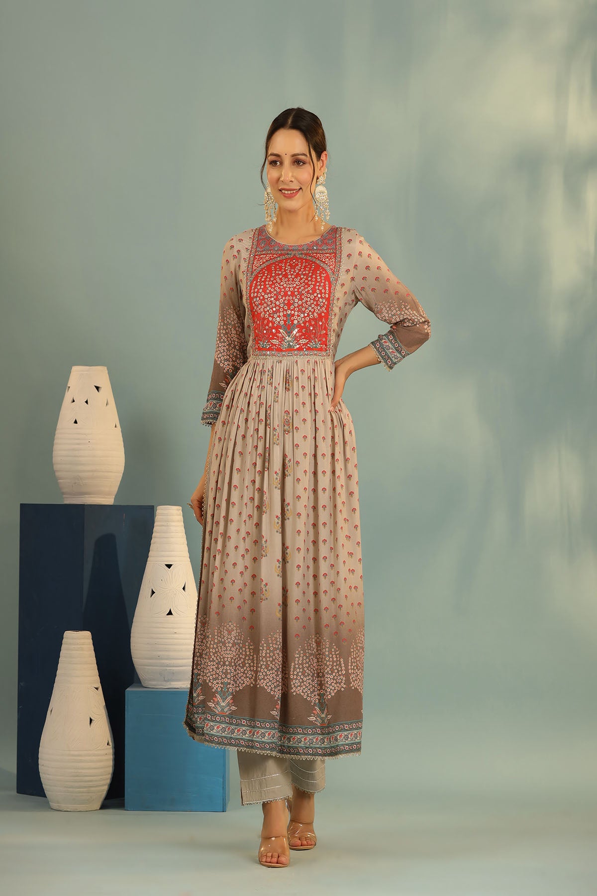 Woman wearing a traditional outfit with red embroidery, standing against a neutral background with decorative vases.