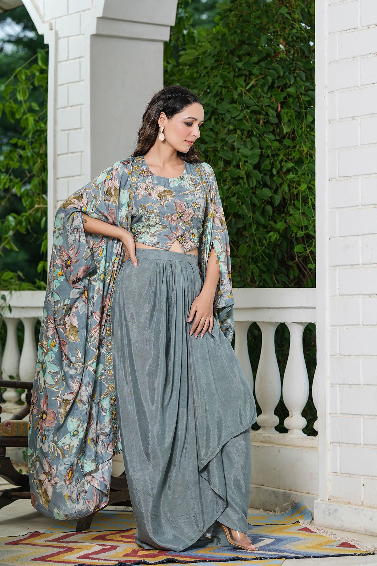 Woman in a floral top and gray skirt standing on a porch with greenery in the background