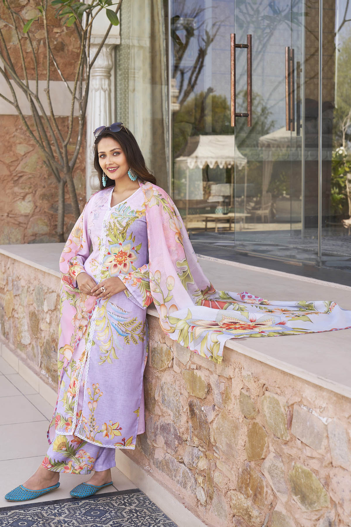 Woman in a floral outfit standing on a stone ledge with a building and garden in the background