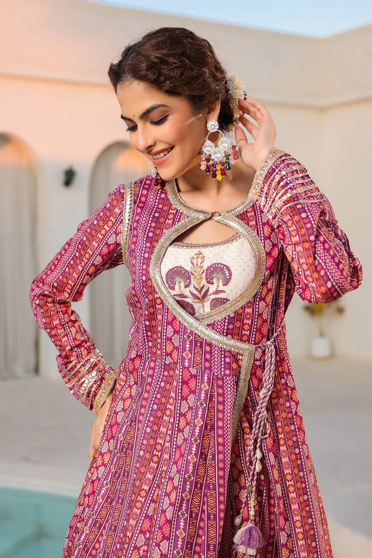 Woman wearing a vibrant pink and red patterned traditional outfit with jewelry, standing against a light background.