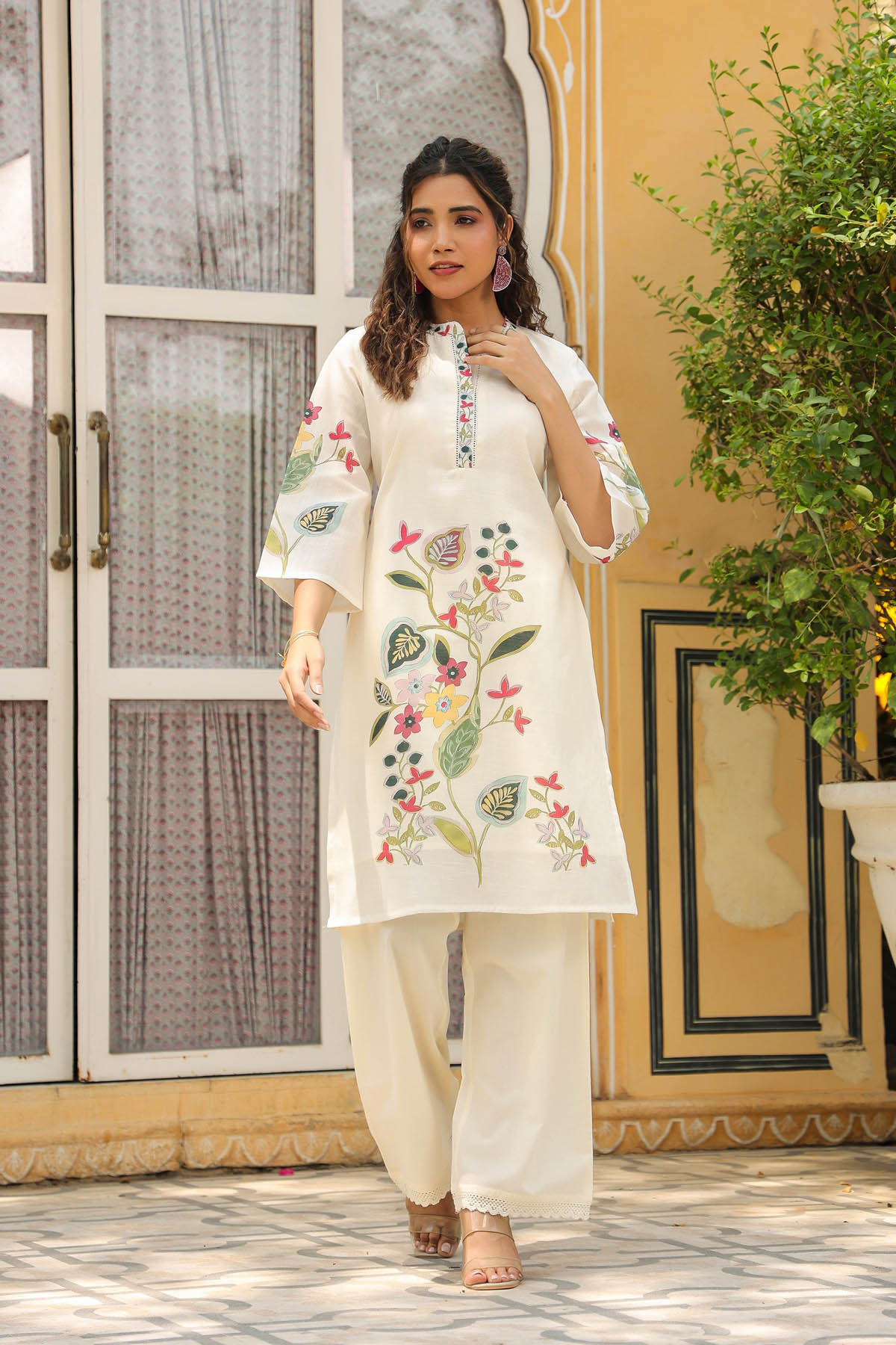Woman wearing a white embroidered outfit standing in a decorative indoor setting.