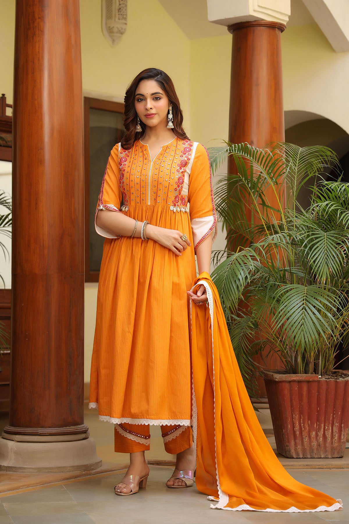Woman in an orange traditional outfit standing in a decorative indoor setting with plants.