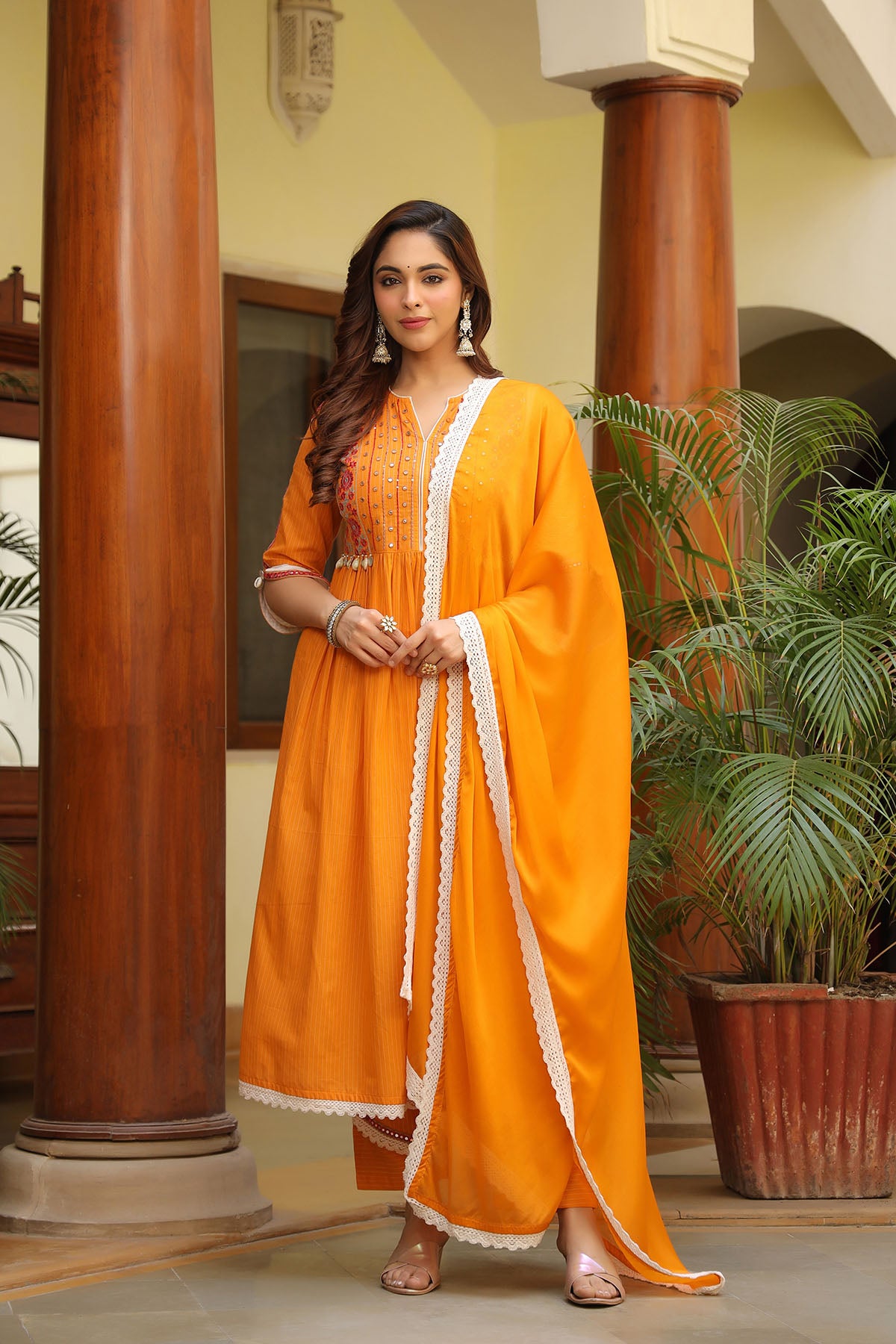 Woman in an orange traditional outfit standing indoors with decorative columns and plants.
