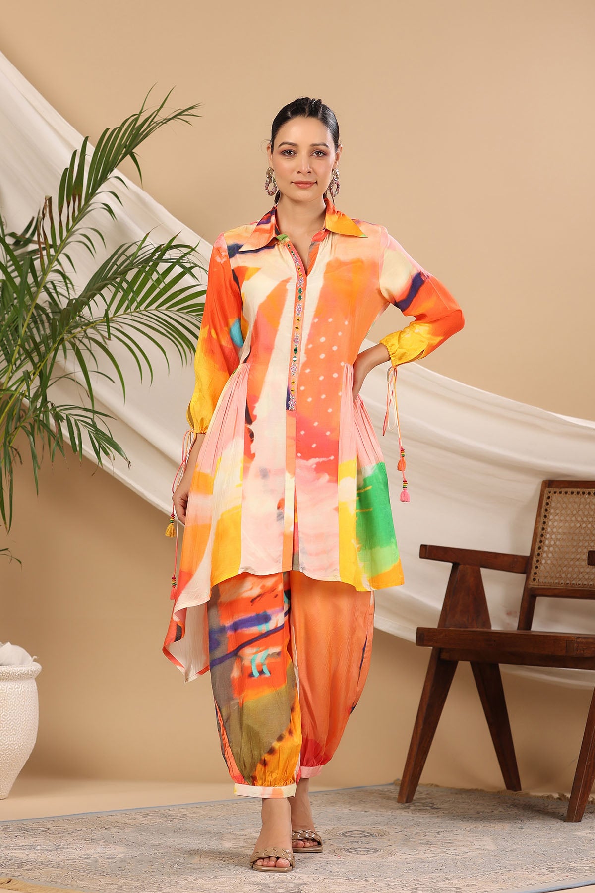 Woman wearing a colorful outfit standing in a room with a plant and chair.