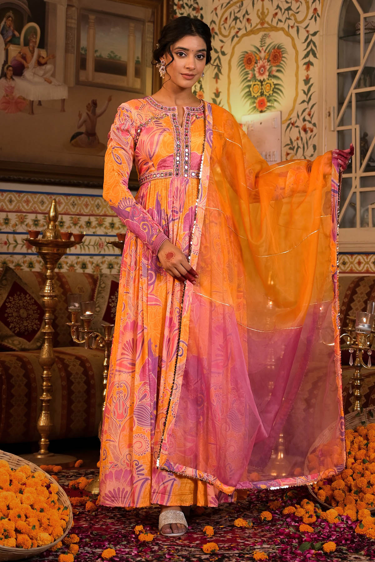 Woman in a vibrant traditional outfit with an orange and pink dupatta in a decorated indoor setting.