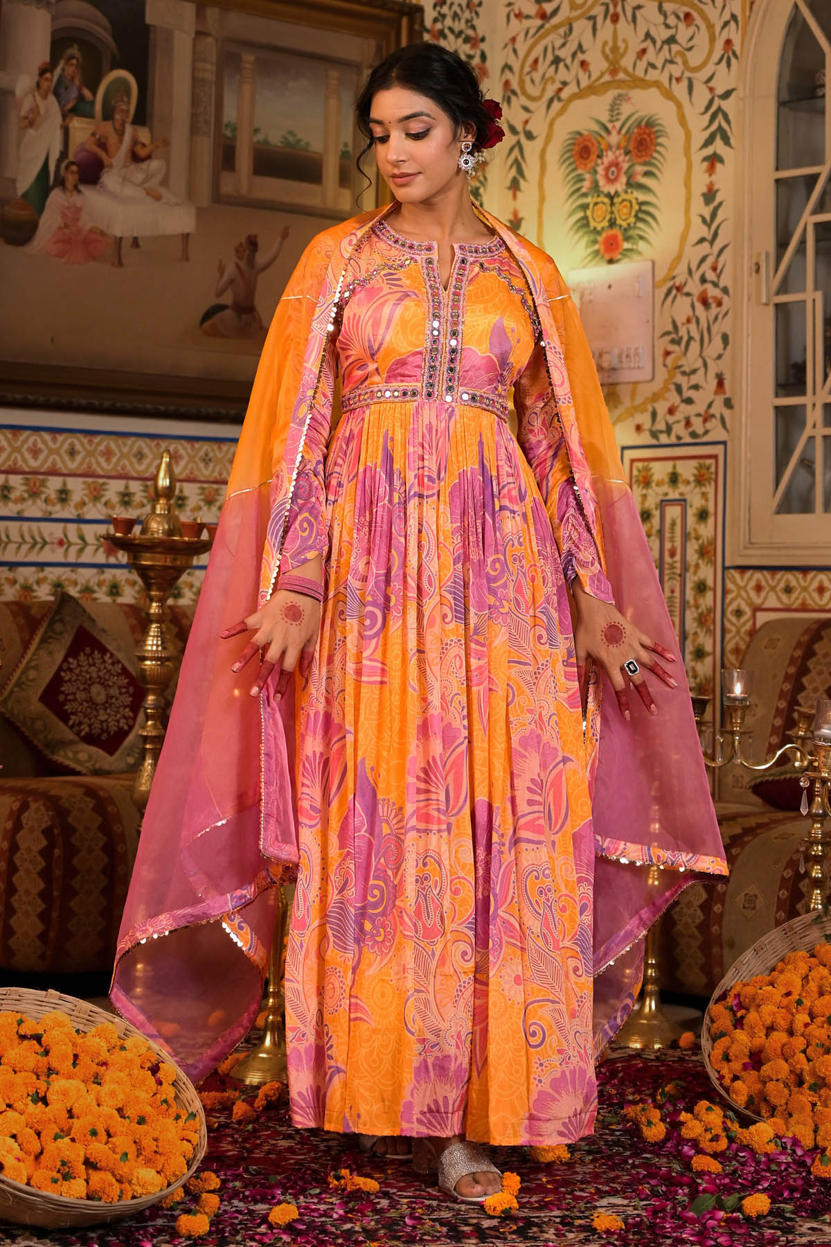 Woman in a traditional orange and pink outfit standing in a decorated room with floral decorations.