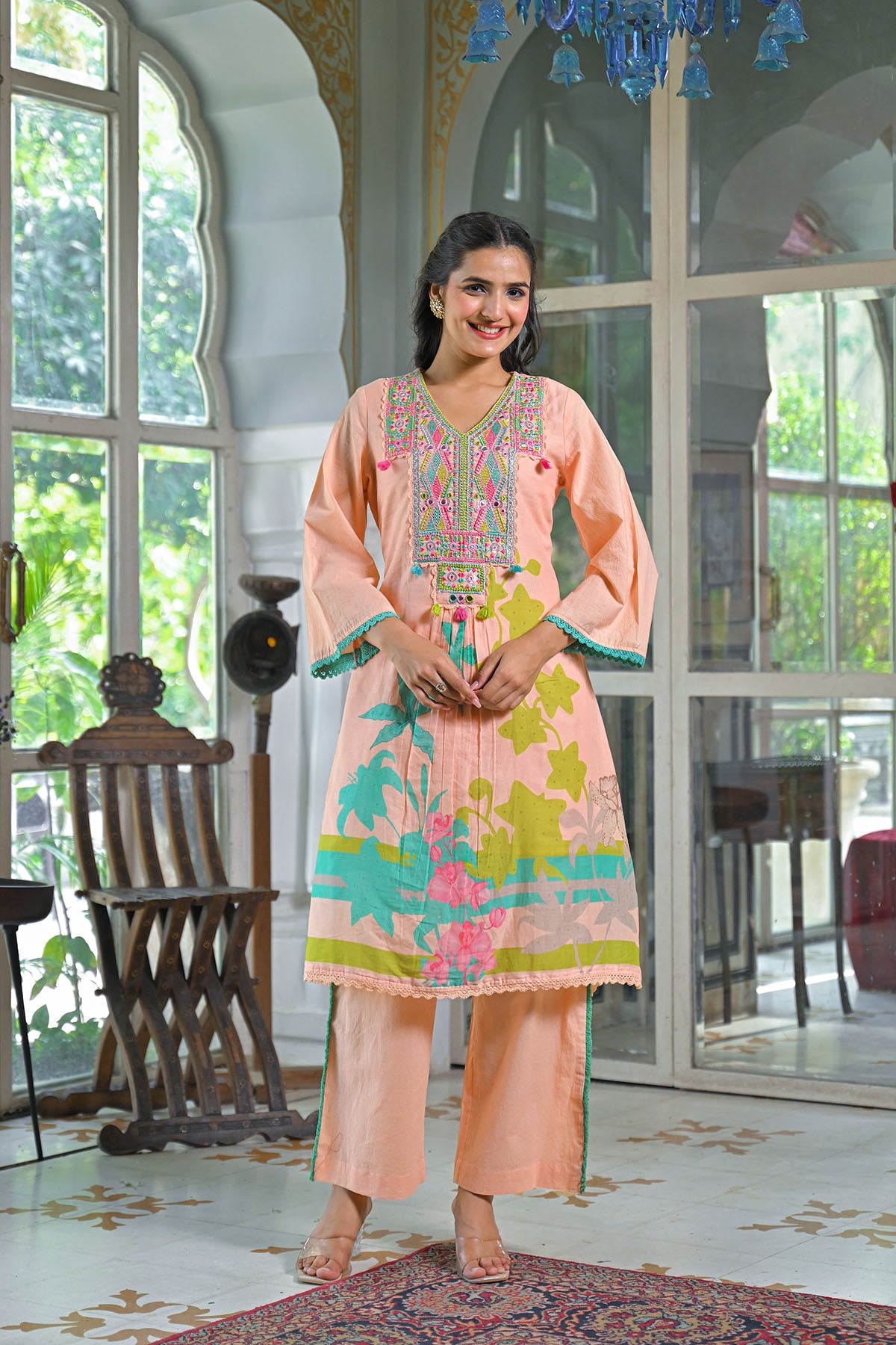 Woman in a peach embroidered outfit standing in a decorated room with large windows.