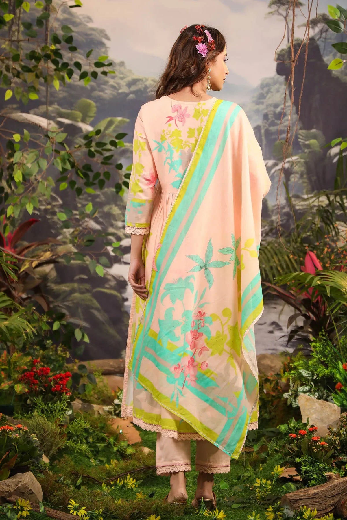 Woman in a floral saree standing in a natural setting with greenery and flowers.
