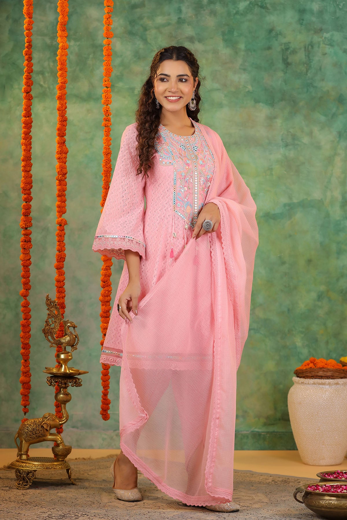 Woman in a pink traditional outfit with embroidery, standing against a decorative background.