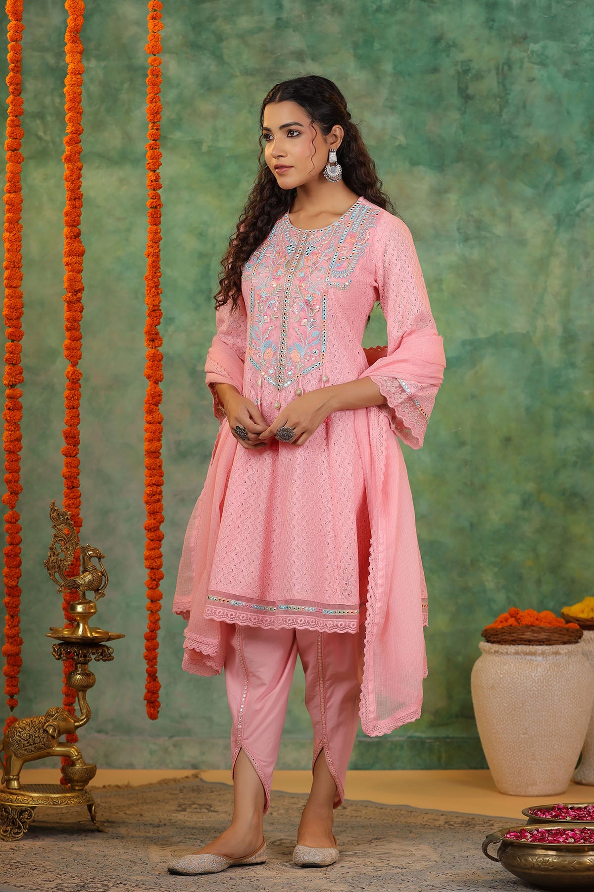 Woman in a pink traditional outfit standing against a textured green wall with decorative elements.