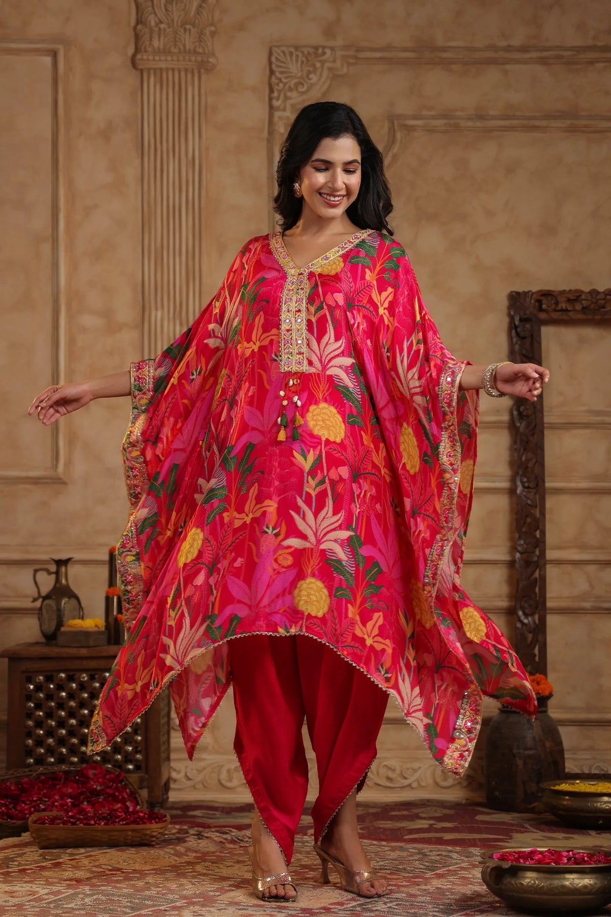 Woman in a vibrant red and pink traditional outfit with floral patterns in an elegant indoor setting.