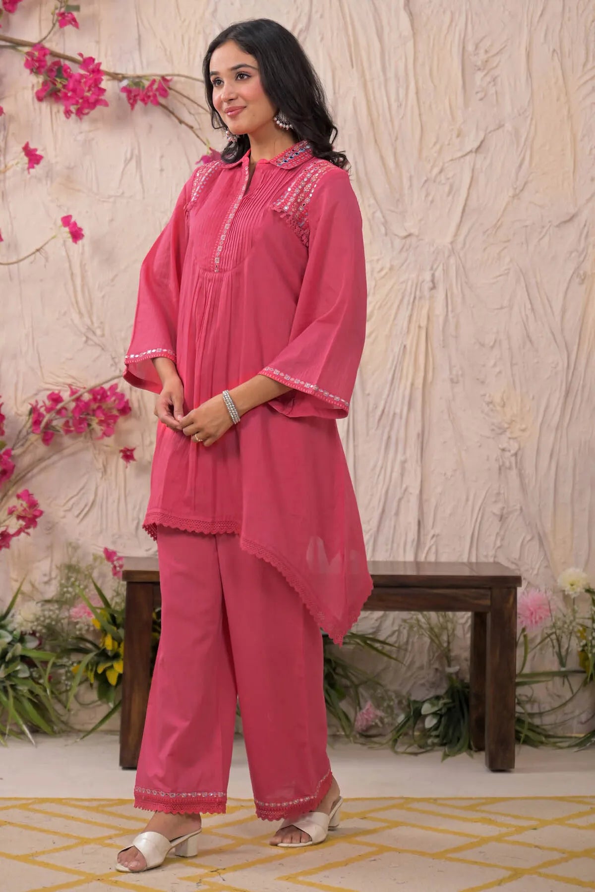Woman wearing a pink traditional outfit with floral decorations against a decorative wall.