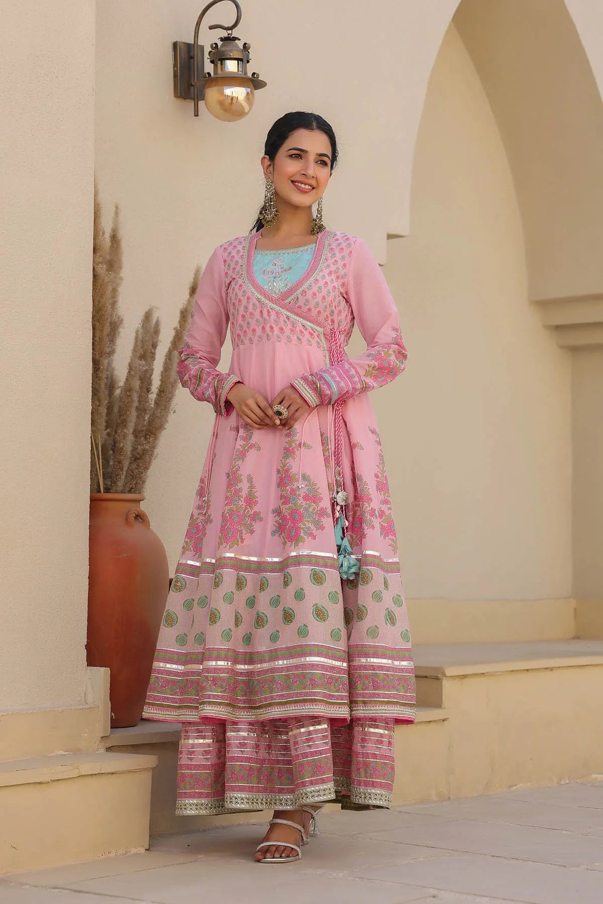 Woman in a pink traditional outfit standing in an outdoor setting with a decorative wall and plant.