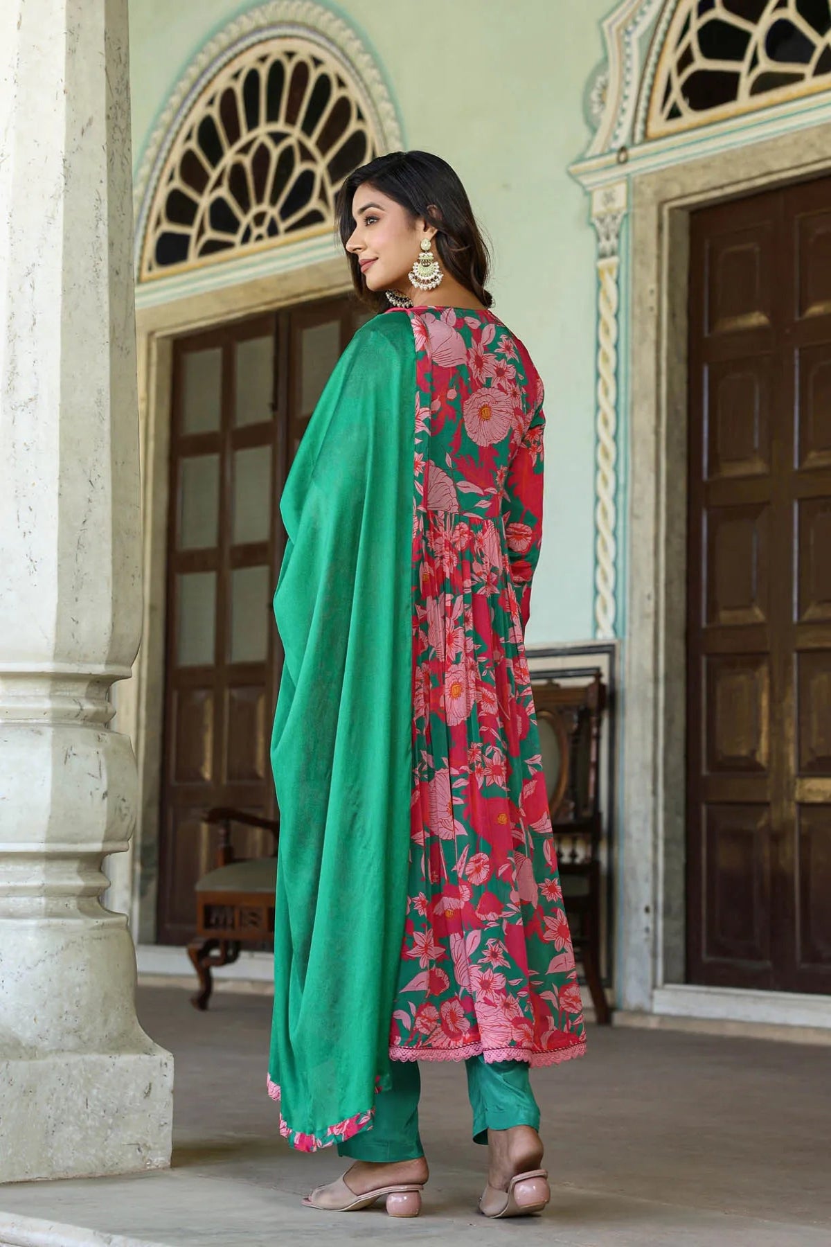 Woman in a floral pink and green outfit standing in an ornate doorway.