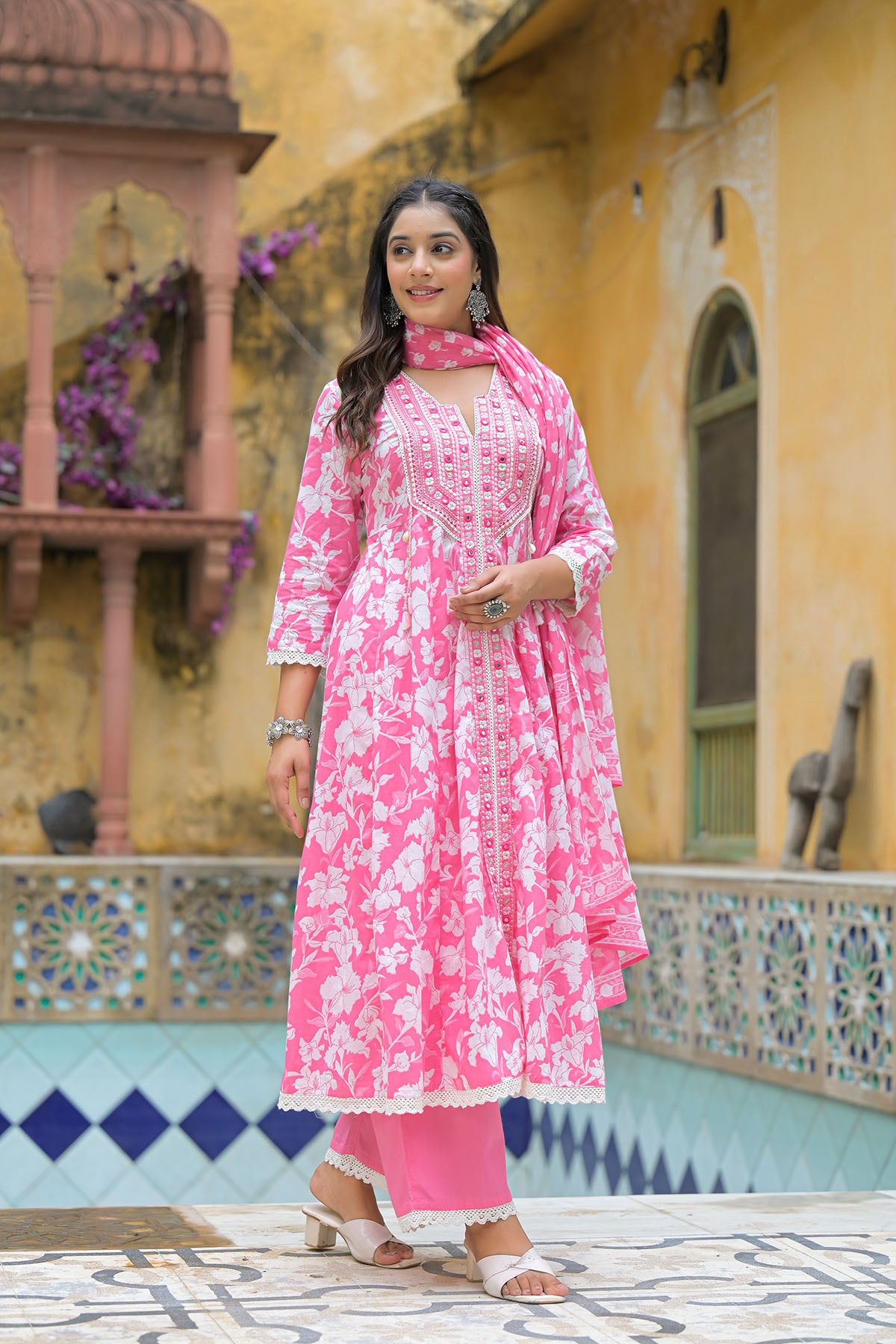 Woman in a pink floral outfit standing in front of a yellow building with decorative elements.