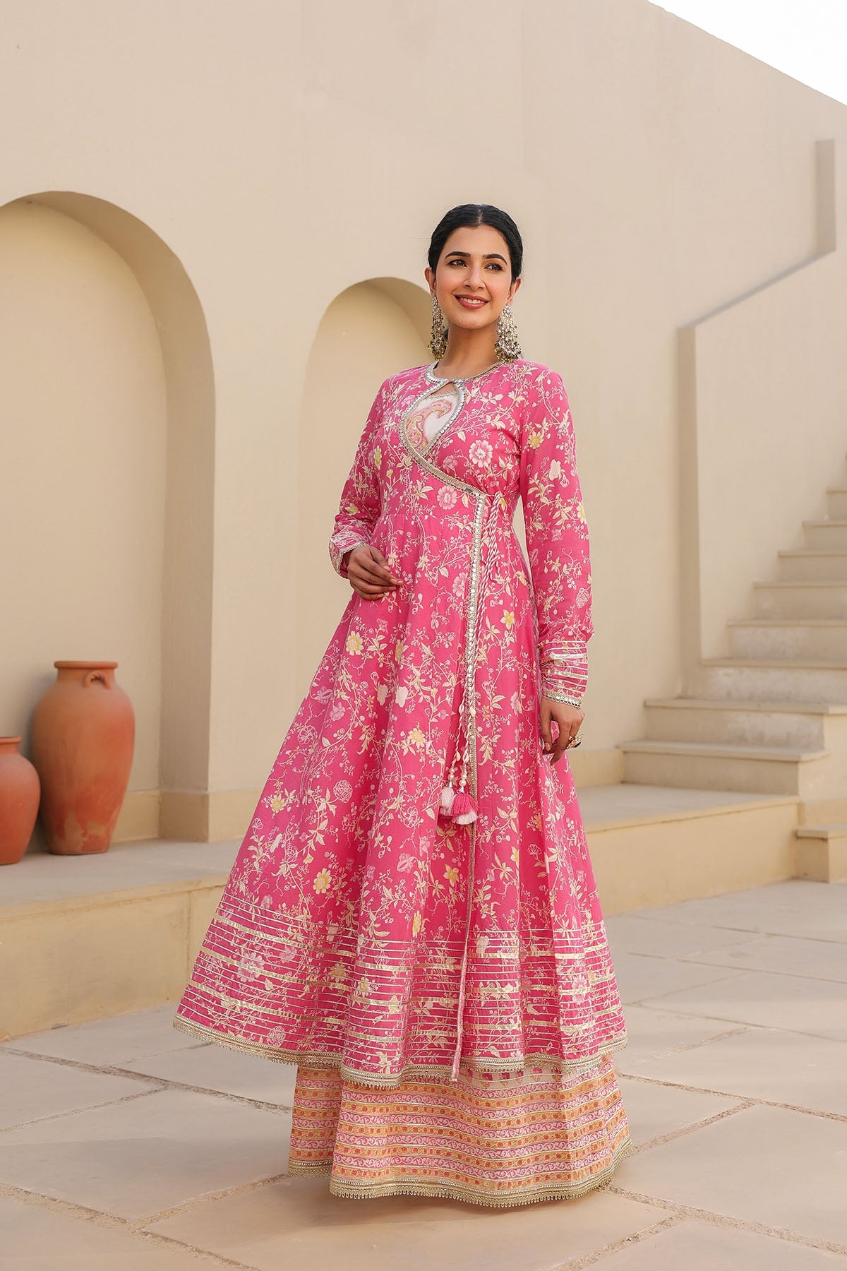 Woman in a pink traditional outfit standing in an outdoor setting with beige walls and steps.