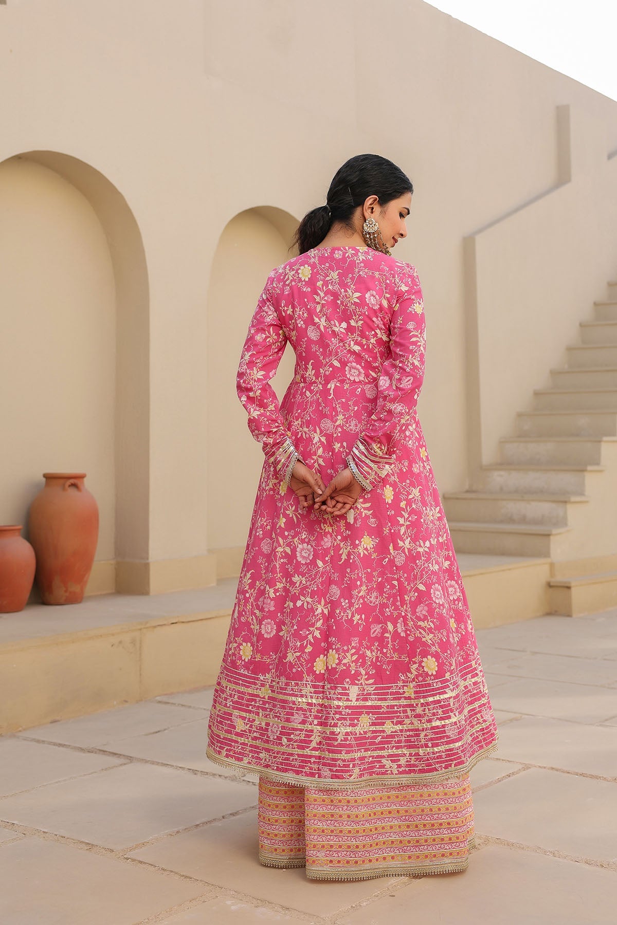 Woman in a pink floral traditional outfit standing in an outdoor setting with arches and steps.