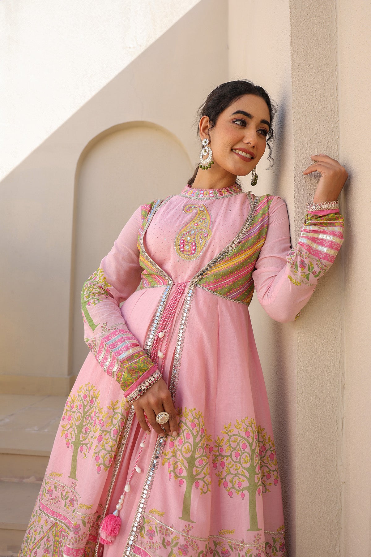 Woman in a pink traditional outfit with intricate patterns standing against a light-colored wall.