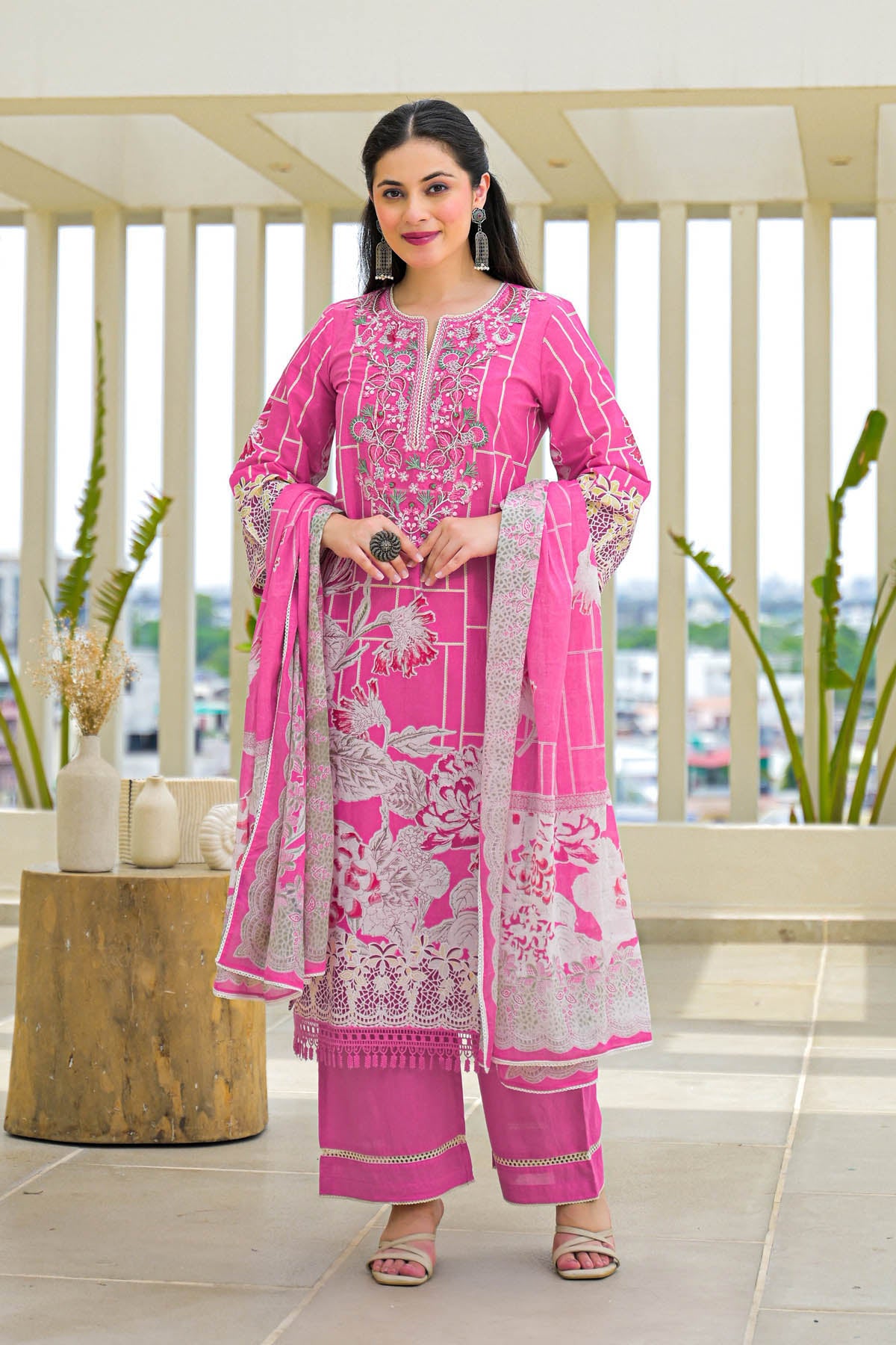 Woman in a pink traditional outfit standing on a balcony with plants and a cityscape view.