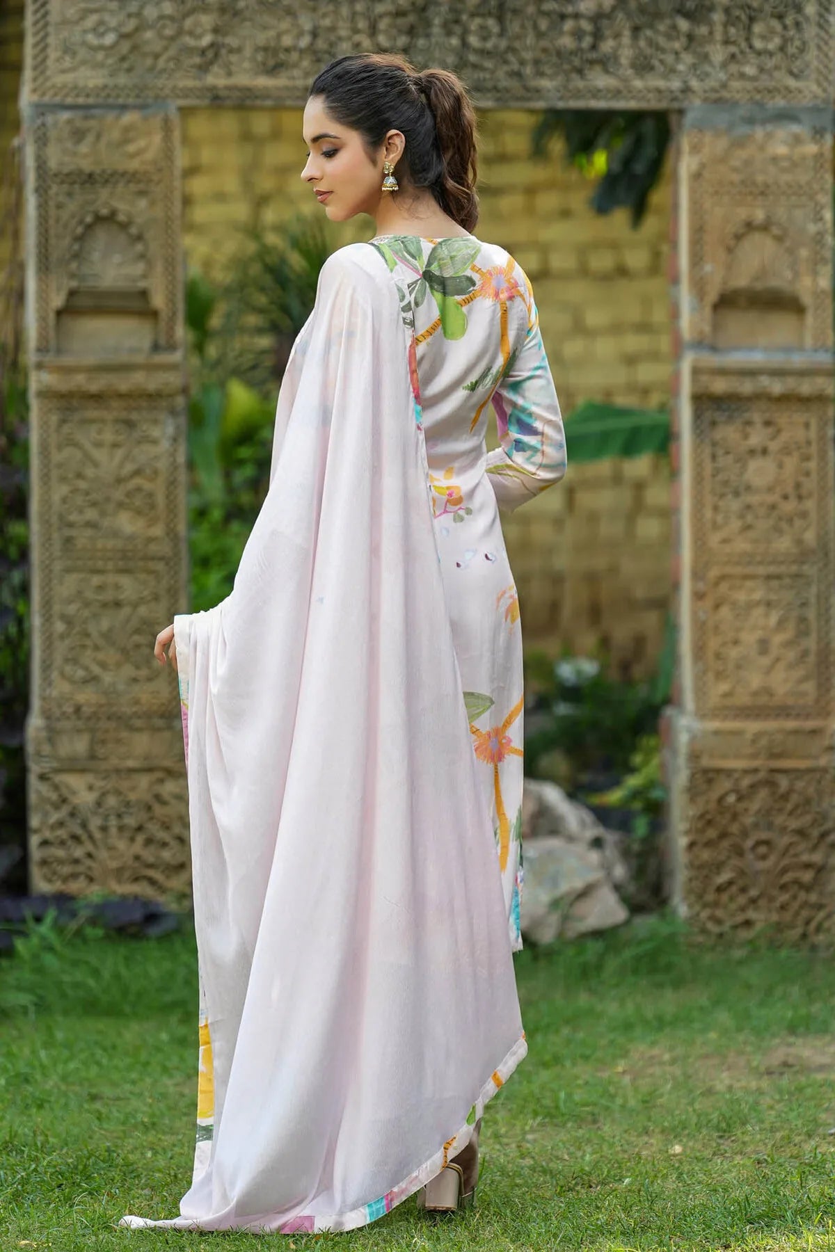 Woman in a white saree with floral patterns standing outdoors near decorative stone walls.