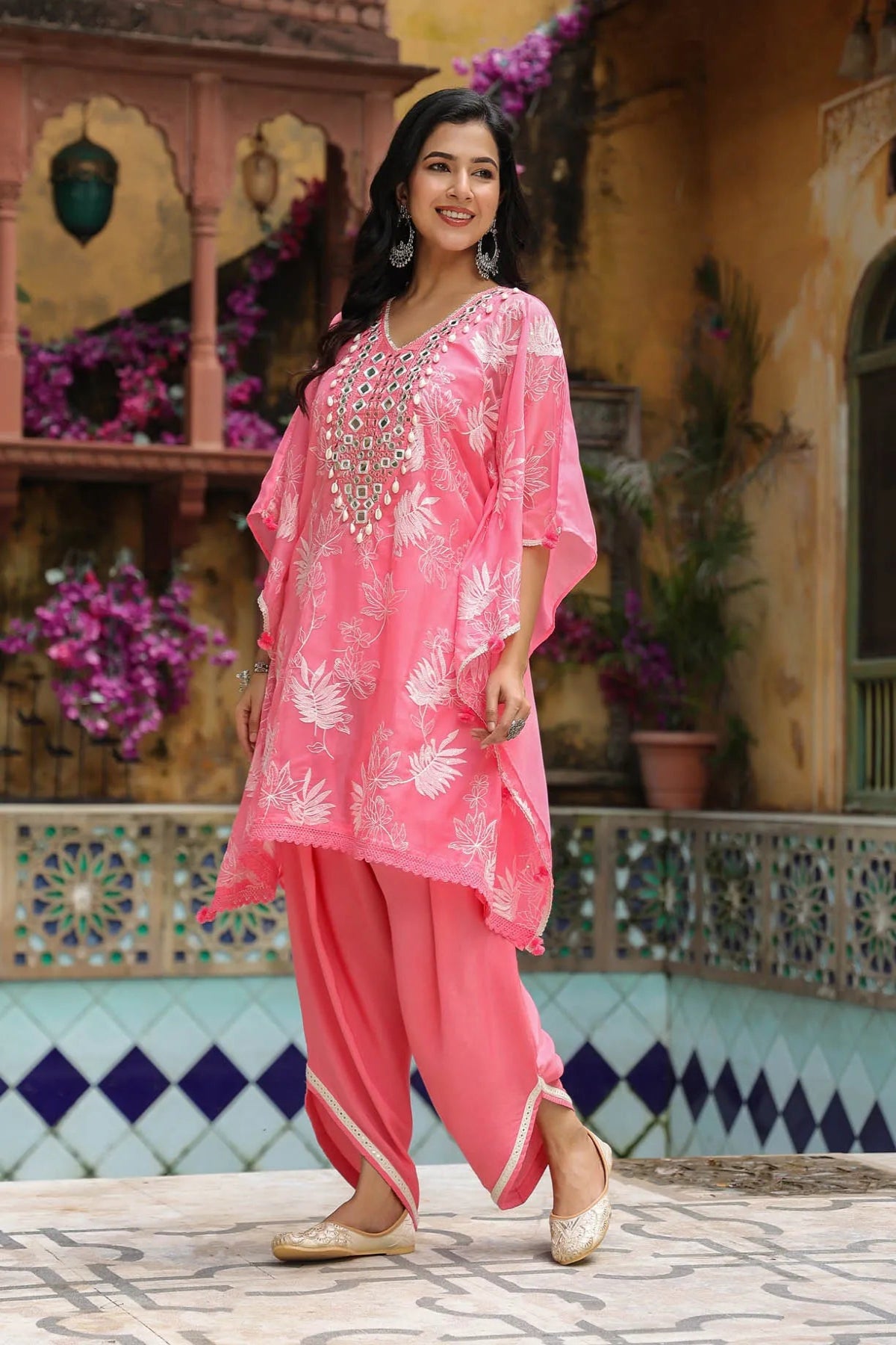 Woman in a pink traditional outfit standing in front of a decorative wall with flowers.