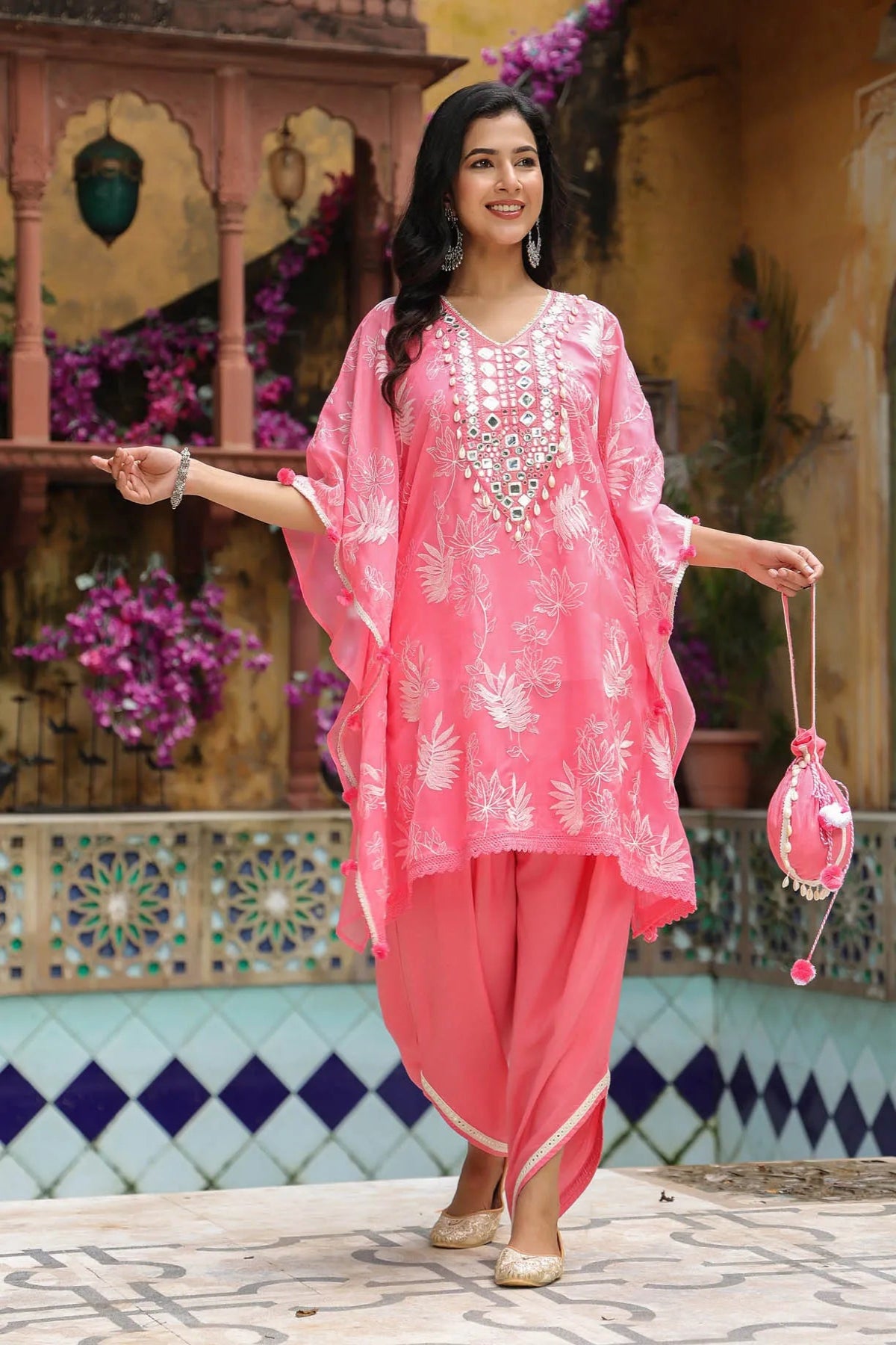 Woman in a pink traditional outfit with white embroidery standing in front of a decorative wall.