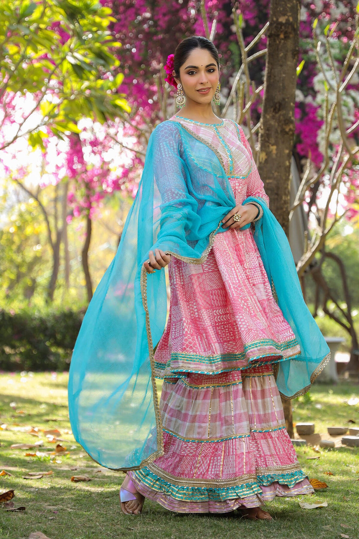 Woman in a traditional pink and blue outfit standing in a garden with pink flowers.