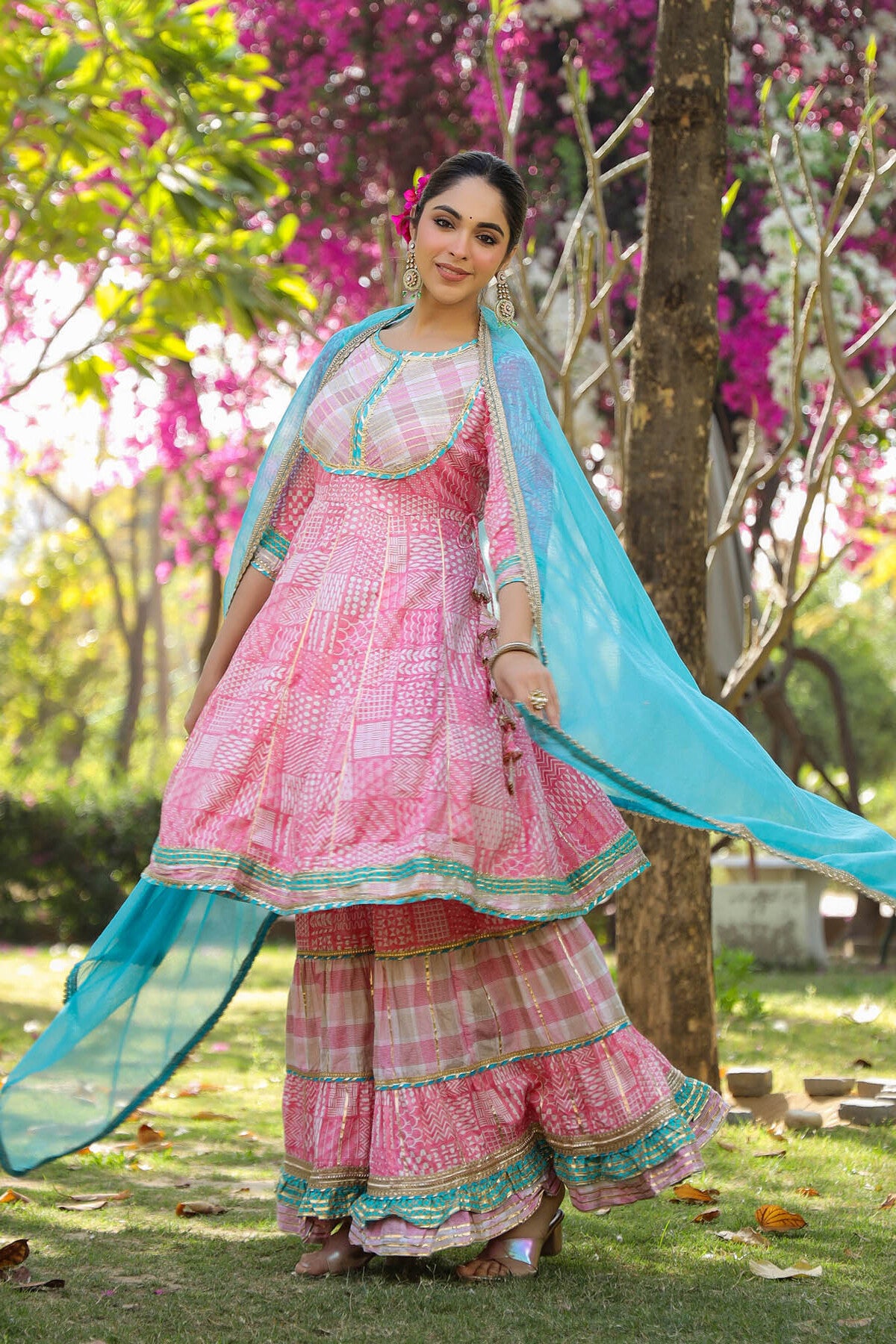 Woman in a pink traditional outfit with a blue dupatta standing under a tree with pink flowers.
