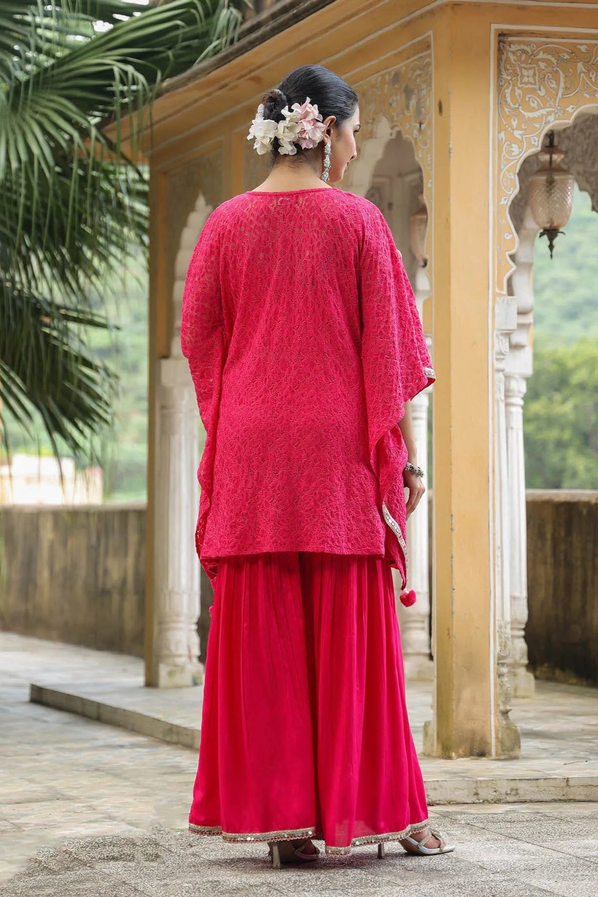 Woman in a red traditional outfit standing in an outdoor setting with greenery.