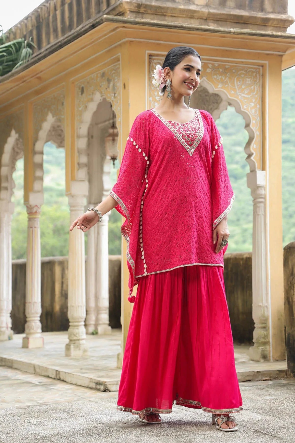 Woman in a pink traditional outfit standing in front of an architectural structure with greenery in the background