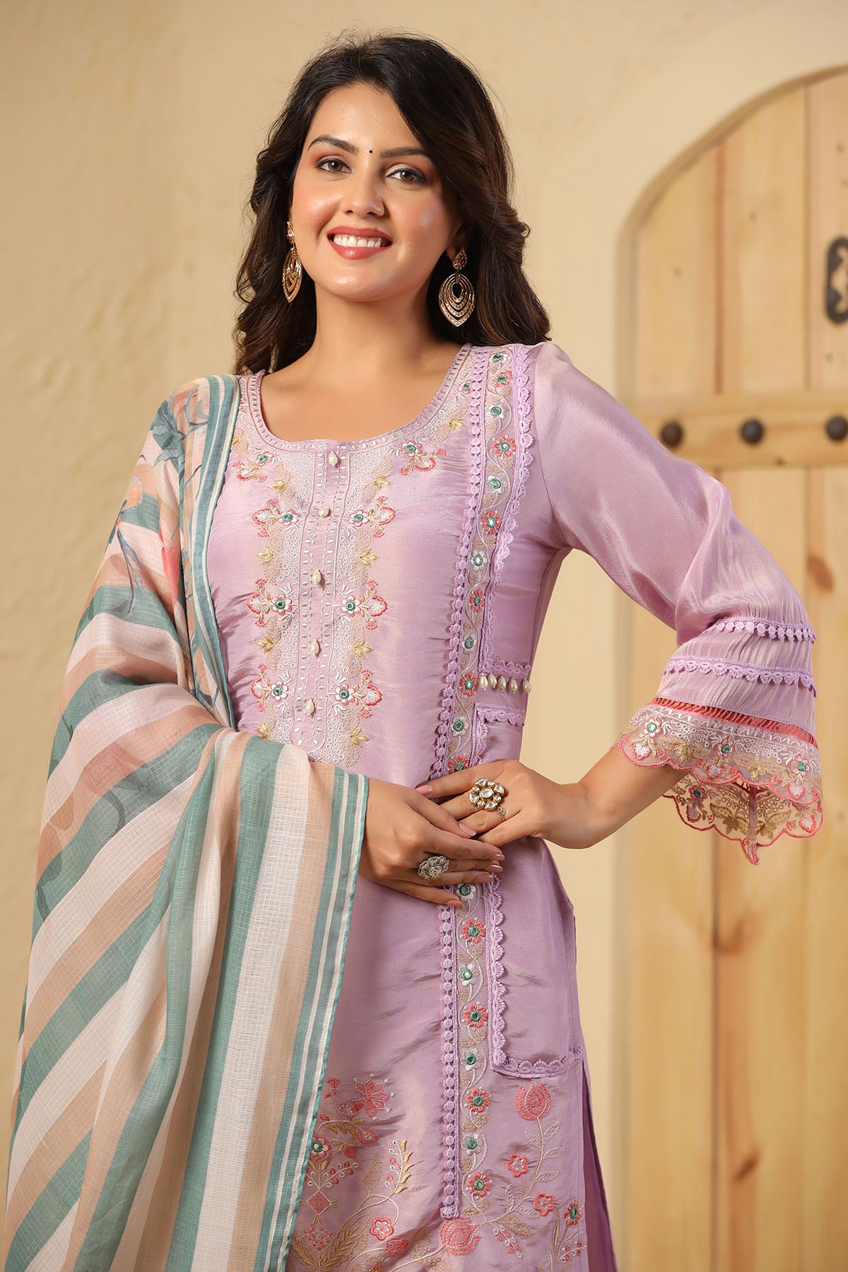 Woman wearing a traditional outfit with a light purple top and colorful dupatta against a wooden door background