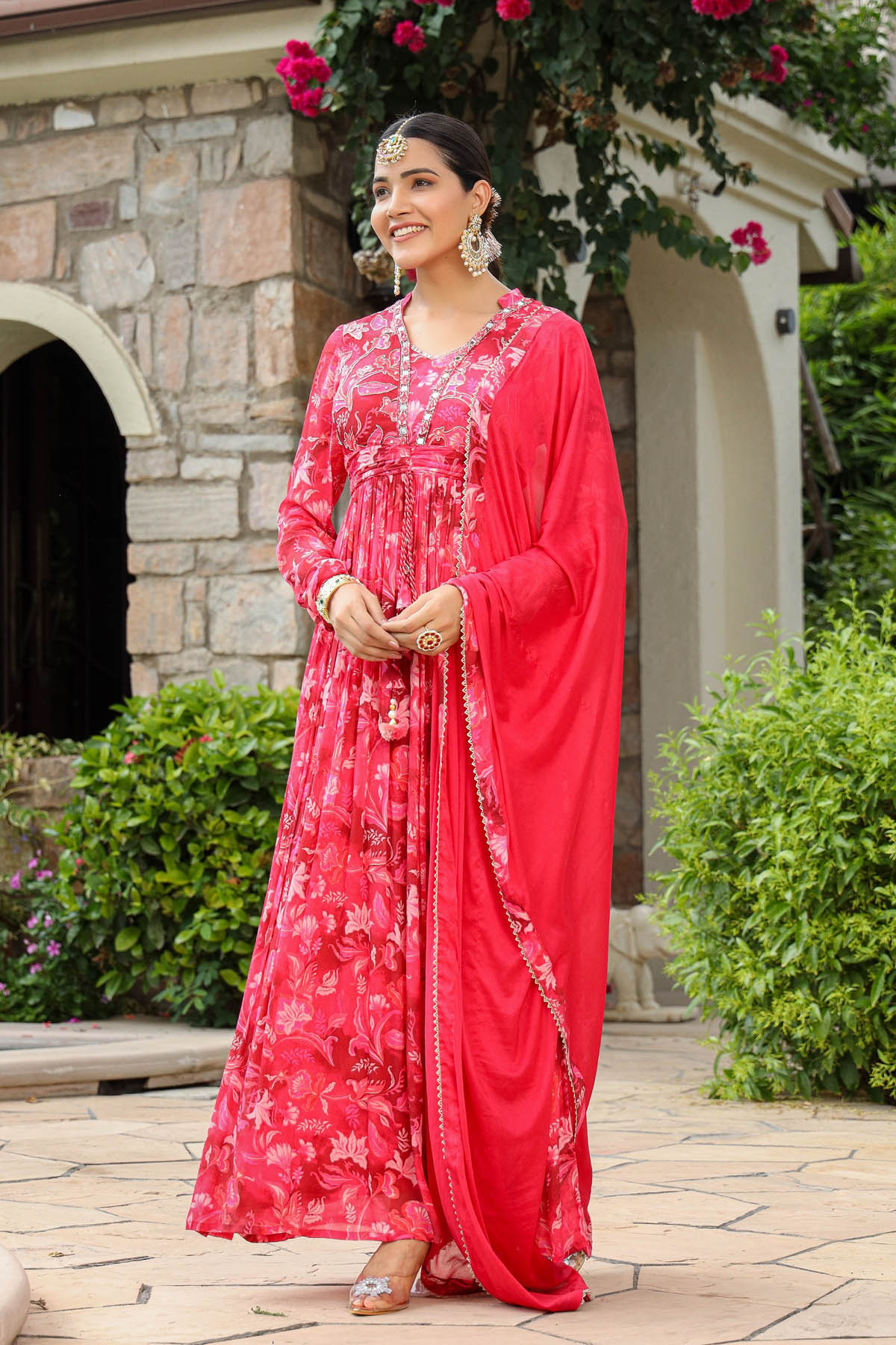 Woman in a red traditional outfit standing outdoors with greenery and a stone building in the background.