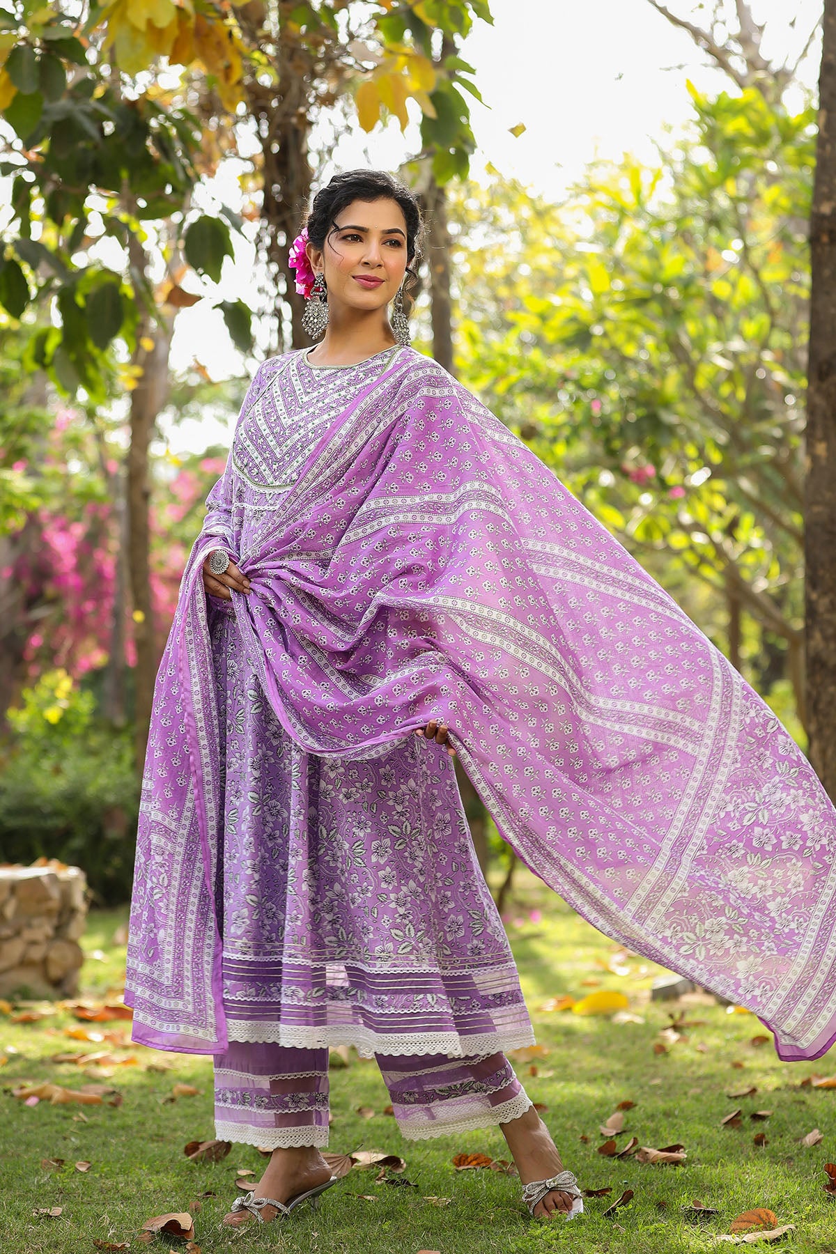 Woman in a purple traditional outfit standing in a garden with greenery and flowers.