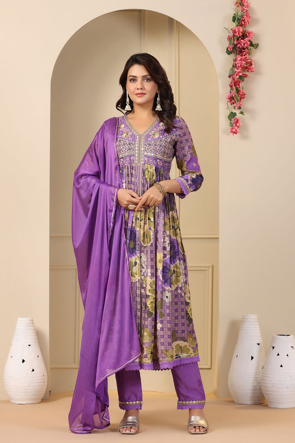 Woman wearing a purple and green traditional outfit with a matching dupatta in a decorated room.