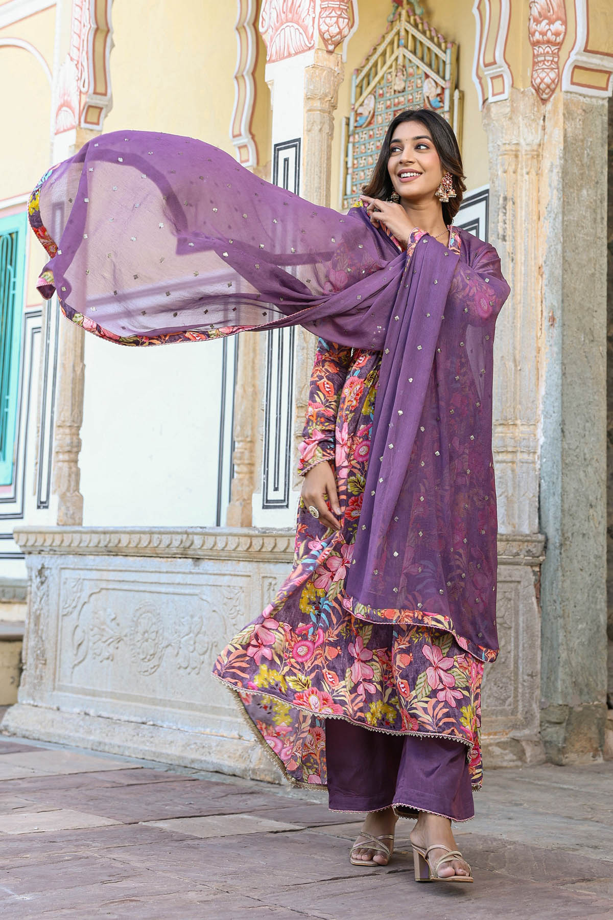 Woman in a traditional outfit with a purple dupatta standing in front of an ornate building.