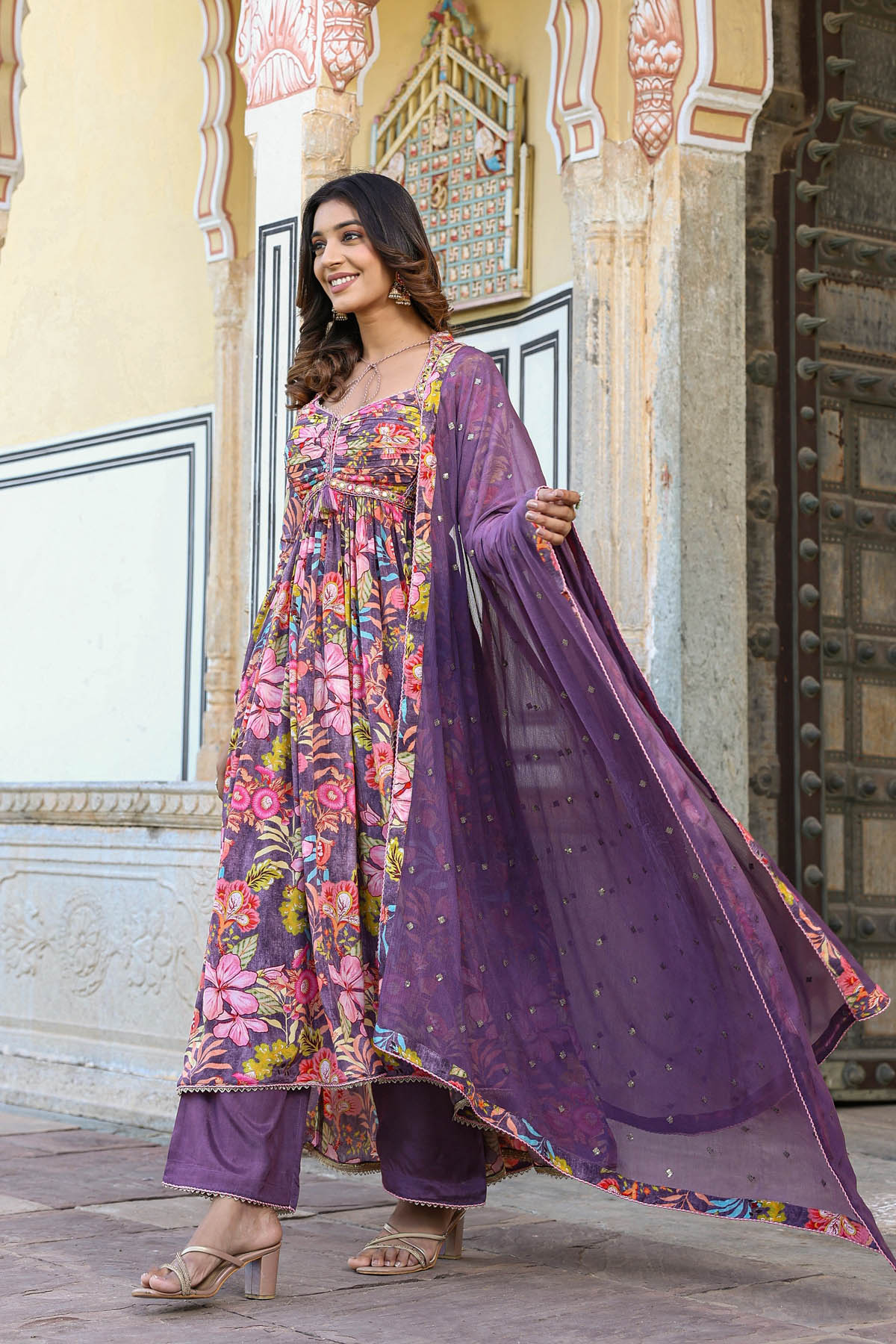 Woman in a traditional outfit with floral and purple design in front of an ornate building.