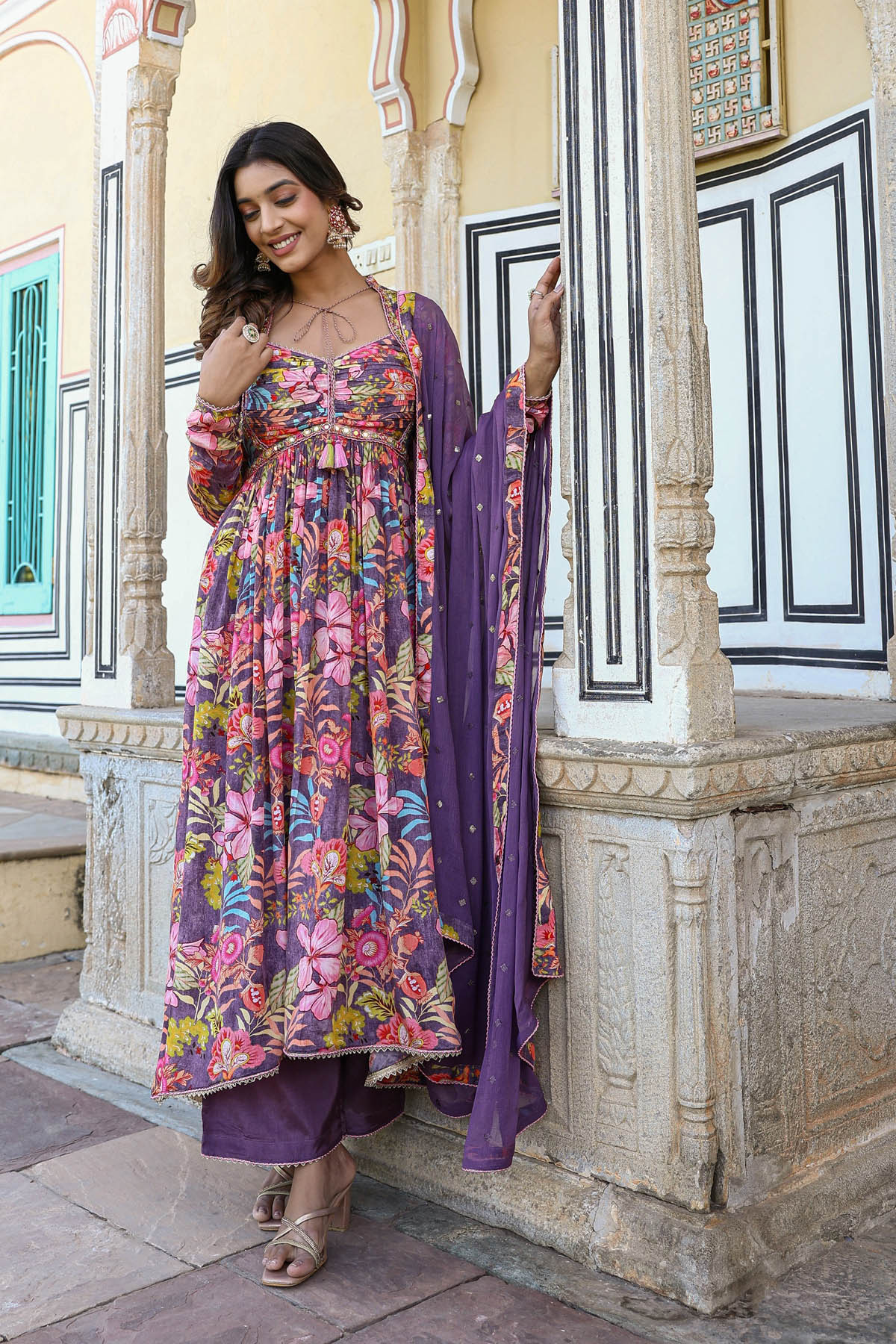 Woman in a colorful floral dress with a purple dupatta standing in front of an ornate architectural background.