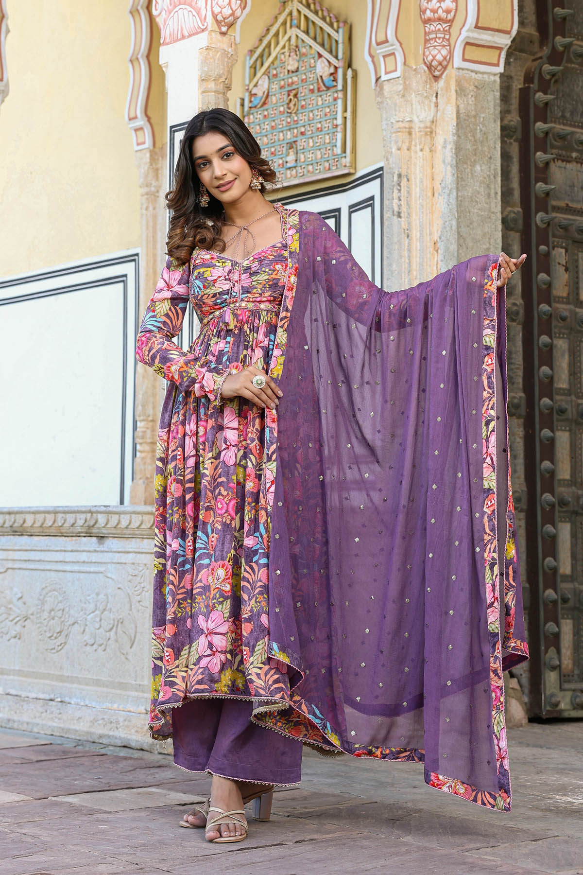 Woman in a colorful traditional outfit with a purple dupatta standing in front of an ornate building.