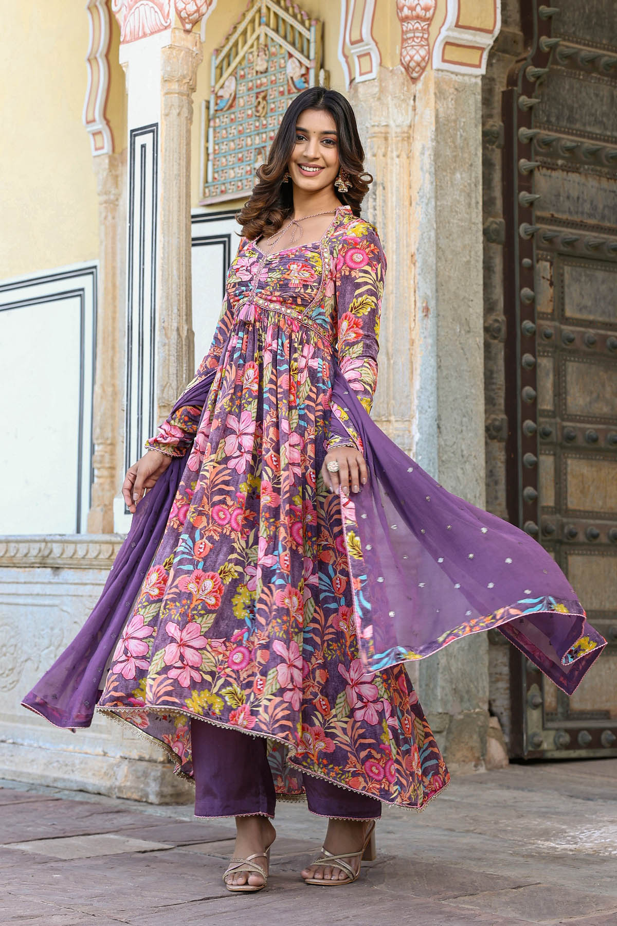 Woman in a colorful floral dress with a purple dupatta standing in front of an ornate building.