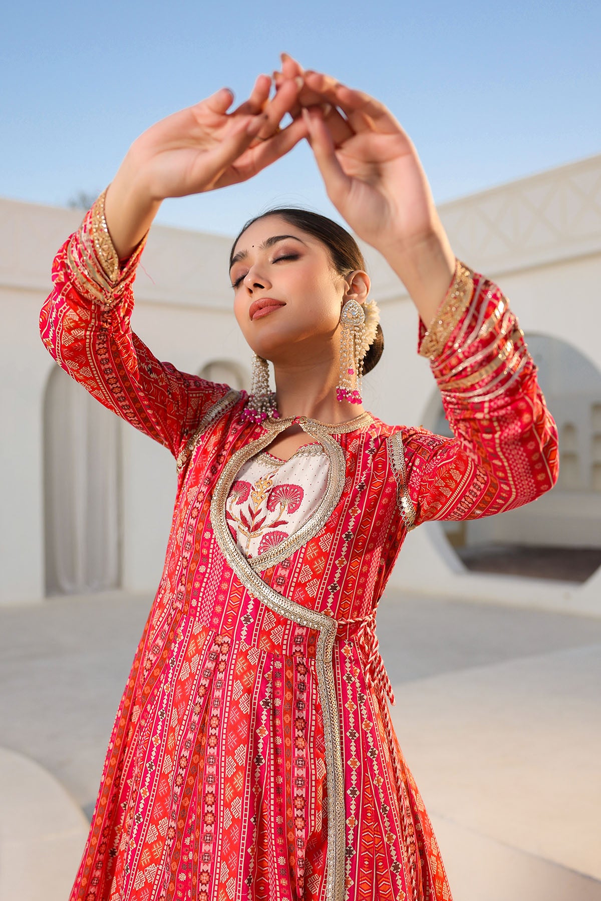 Woman in a red traditional outfit with intricate patterns, standing against a light-colored building.