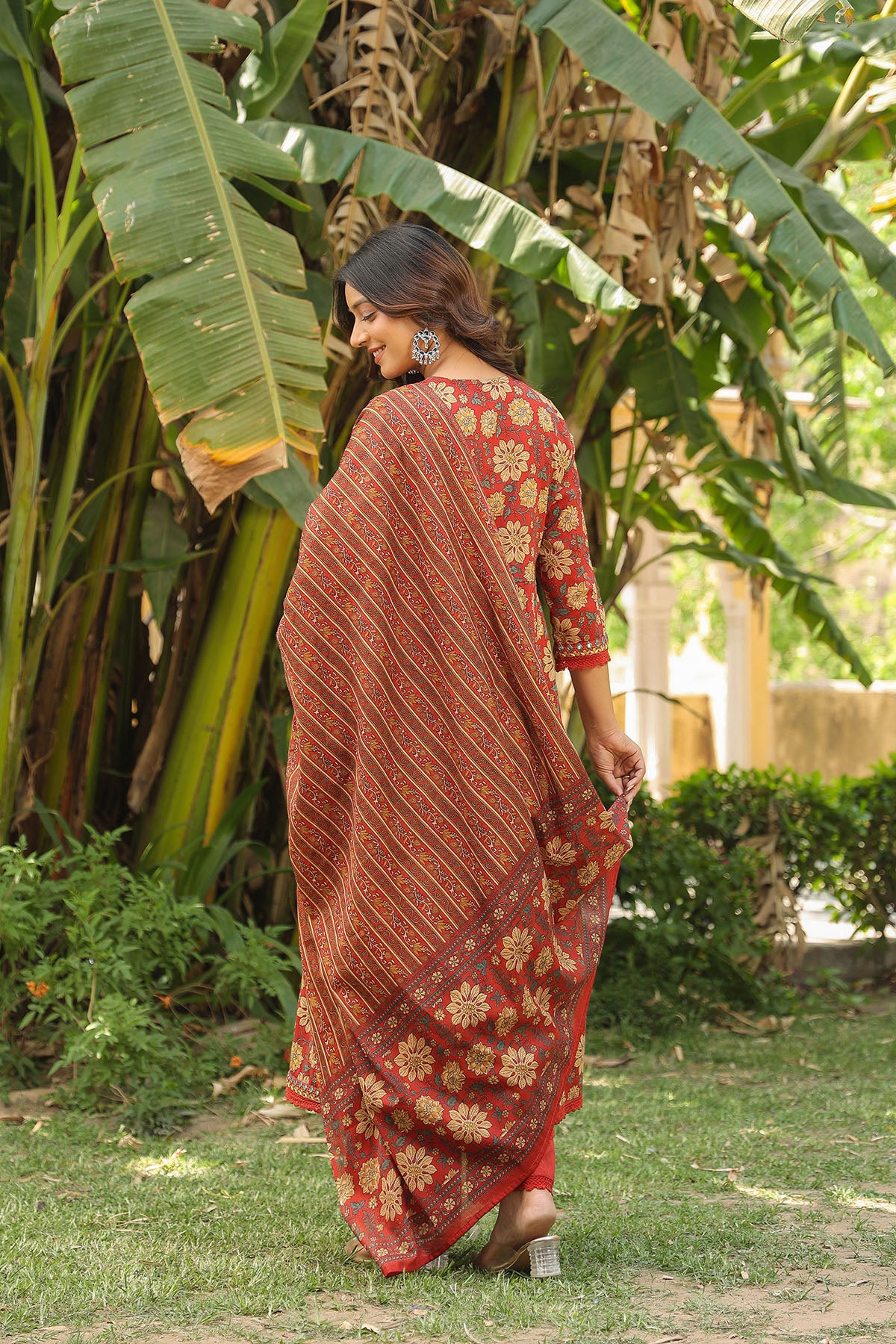 Woman in a red and gold saree walking outdoors among green plants