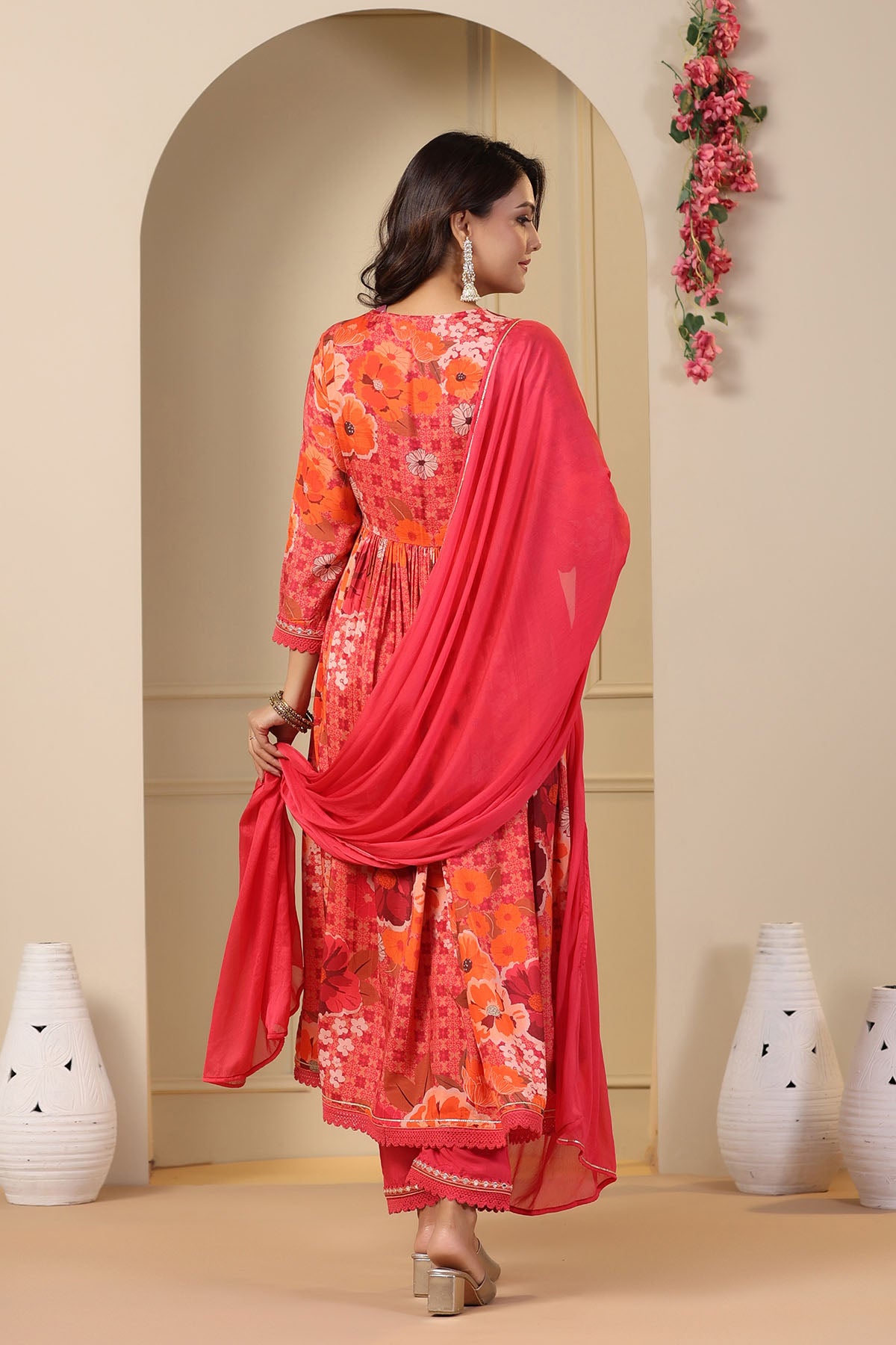 Woman in a red and orange traditional outfit with a pink dupatta standing in a decorated room.