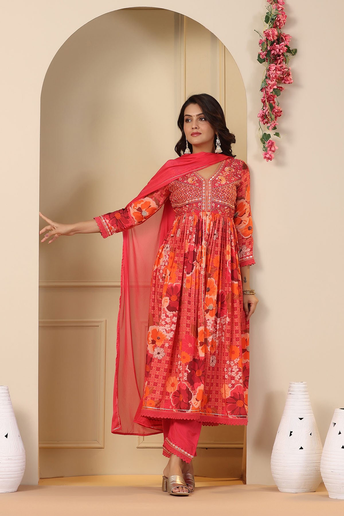 Woman in a red and pink traditional outfit standing in a decorated room with vases.
