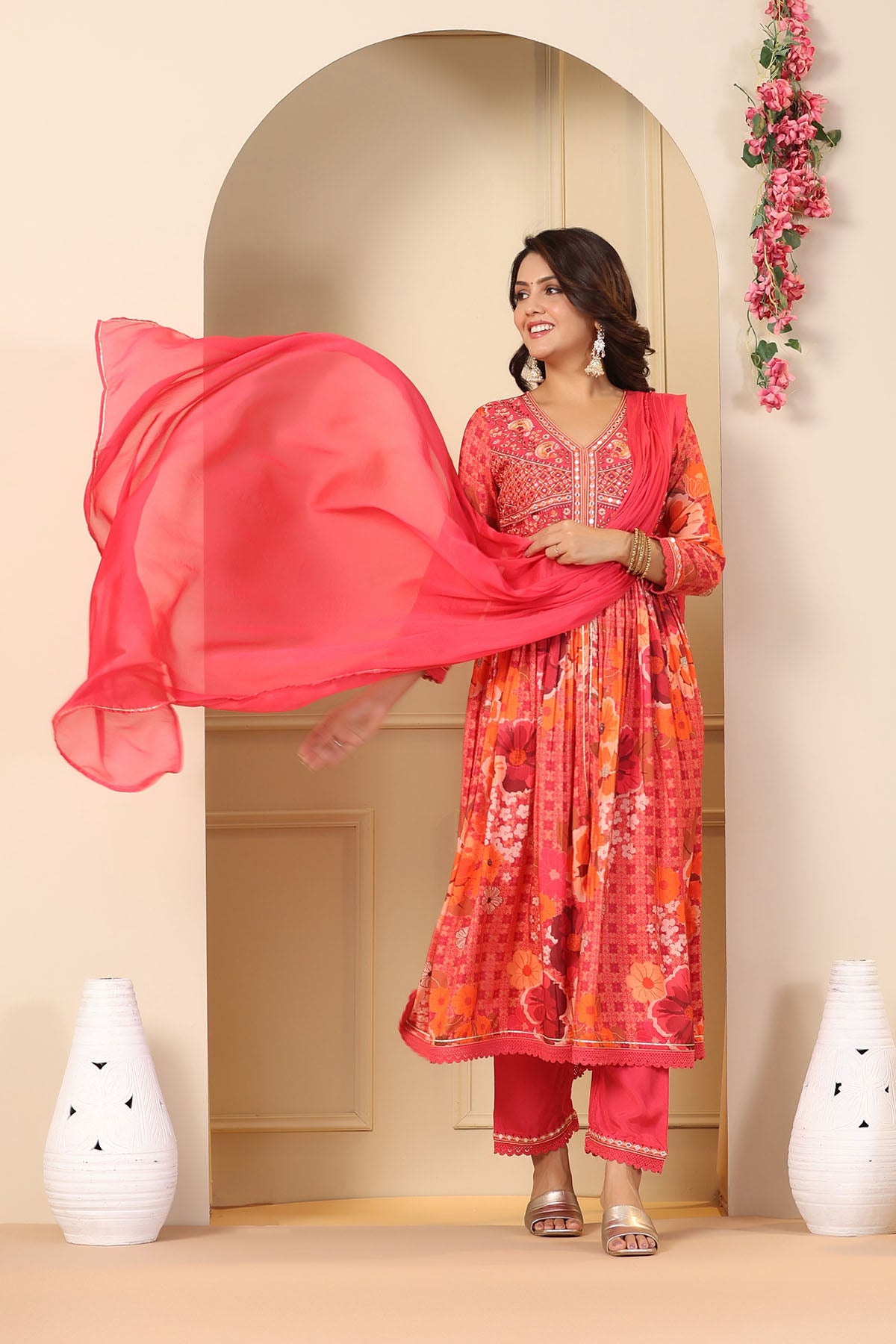 Woman in a red and orange traditional outfit holding a pink dupatta in a decorated room.